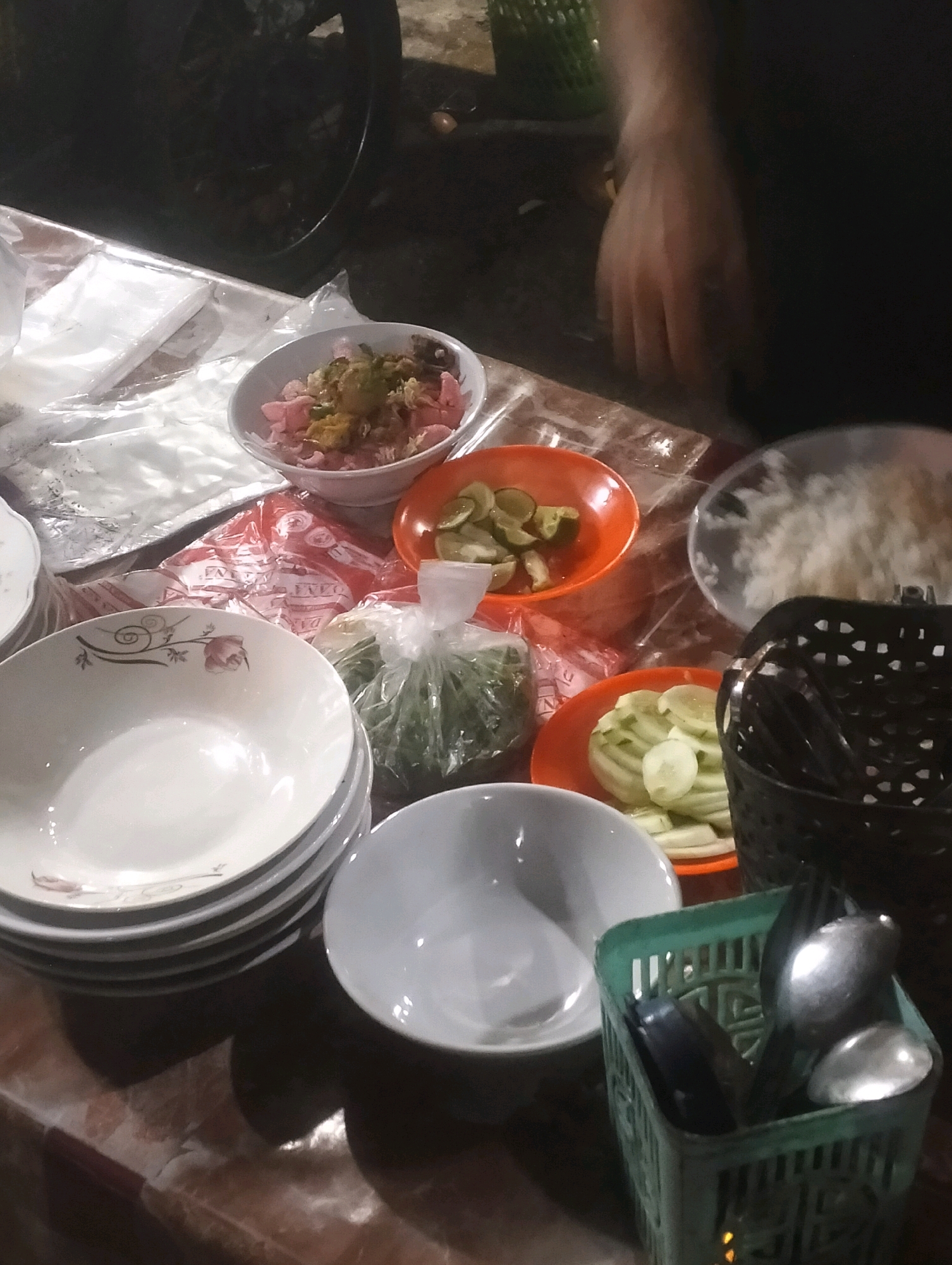 An overhead shot captures a dimly lit outdoor food stall in Air Jamban, Indonesia, featuring various dishes and utensils laid out on a patterned marble table. In the foreground, a stack of white ceramic bowls with a floral design sits to the left, while a single white bowl is placed to its right. A green plastic basket filled with silverware is positioned on the right, next to a black basket also containing utensils. Further back, an orange bowl holds sliced cucumbers and lime wedges, and another orange bowl contains sliced cucumbers. A clear plastic bag filled with green leafy vegetables is visible in the mid-ground, alongside a bowl of what appears to be rice. To the left of the food is a stack of white paper napkins. A blurred hand is visible in the upper right corner, suggesting someone is preparing or serving the food. The background is dark, with a glimpse of a dark circular object, possibly a wheel or pot, and a green woven basket.