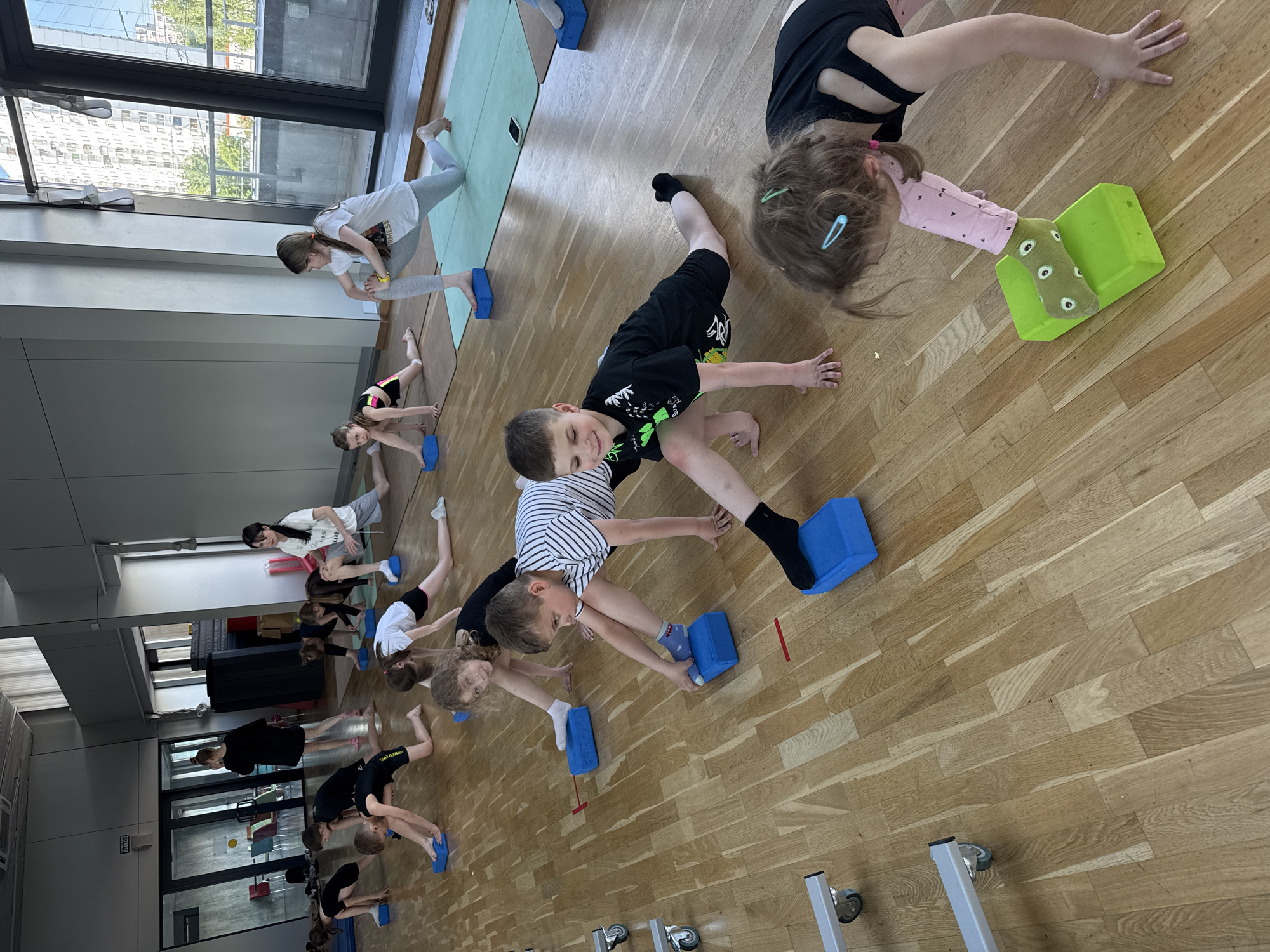 This image captures an indoor physical activity session, likely a gymnastics or stretching class, featuring a group of children and some adults. The setting is a brightly lit room with a light-colored wooden floor and large windows that offer a view of city buildings, suggesting an urban environment such as Khimki, Russia.
The participants are engaged in various exercises, primarily involving stretches and balance poses. Many children are positioned in a lunge-like stance, using colorful foam blocks (mostly blue, with one green) for support under their feet or hands. Some are also seen in plank-like or kneeling positions. Their expressions range from focused concentration to slight smiles, indicating engagement in the activity. Most individuals are dressed in athletic wear, including t-shirts, shorts, leggings, and socks.
Notable details include:
* **Props:** Numerous blue and one bright green foam blocks are scattered across the floor, used by the participants for their exercises.
* **Mats:** Green exercise mats are visible on the floor near the windows.
* **Lighting:** The room is well-lit by natural light streaming through the large windows, suggesting it is daytime.
* **Background:** Beyond the windows, multi-story urban buildings are visible. Inside the room, there are also reflective surfaces, possibly mirrors or glass partitions, and some furniture on wheels.
* **Visible Text:** The word "ВЫХОД" (meaning "EXIT" in Russian) is visible on a door in the mid-left background. A girl's t-shirt in the background has "ARMEES" printed on it.