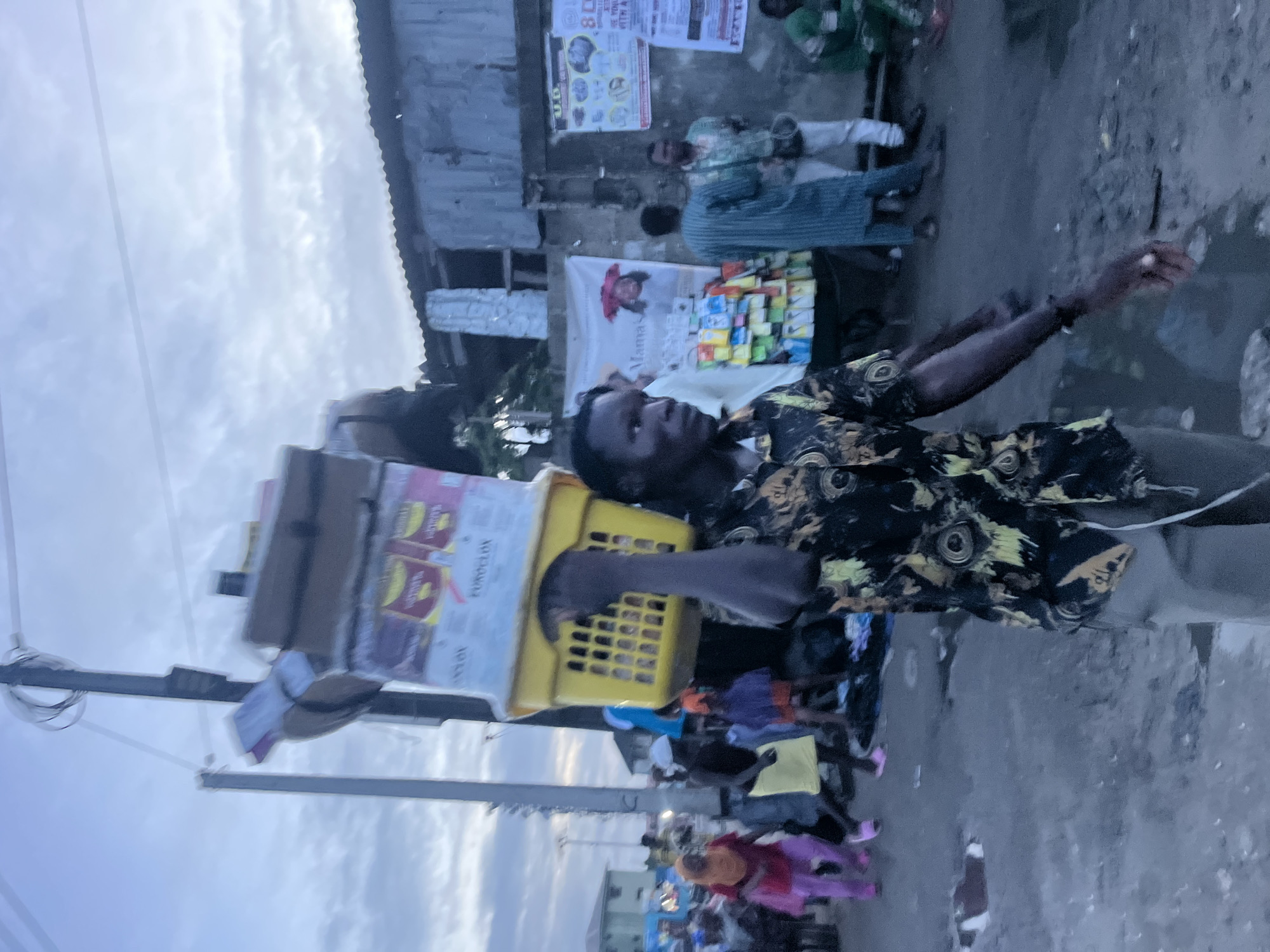 The image depicts a street scene in Ado, Nigeria, likely in a bustling market area given the presence of vendors and the general atmosphere. A man in the foreground is carrying a large stack of goods on his head, balanced on a yellow plastic basket. The goods appear to be packaged items, possibly food or household products, with some text visible on the packaging. He is wearing a patterned shirt and light-colored pants.
In the background, other individuals are visible, some shopping or walking. A woman in a red hijab and pink pants is to the left, and a person in a yellow skirt is also present. A vendor stall with various items, possibly phone accessories or small goods, is set up against a wall. A banner with text and an image is hanging behind the stall. The architecture in the background suggests a mix of buildings, some appearing to be made of corrugated metal. The ground is a dirt or paved surface with some puddles, indicating it might have recently rained. The sky is overcast with clouds, suggesting either early morning or late afternoon lighting. The overall impression is one of daily life and commerce in a busy urban environment.