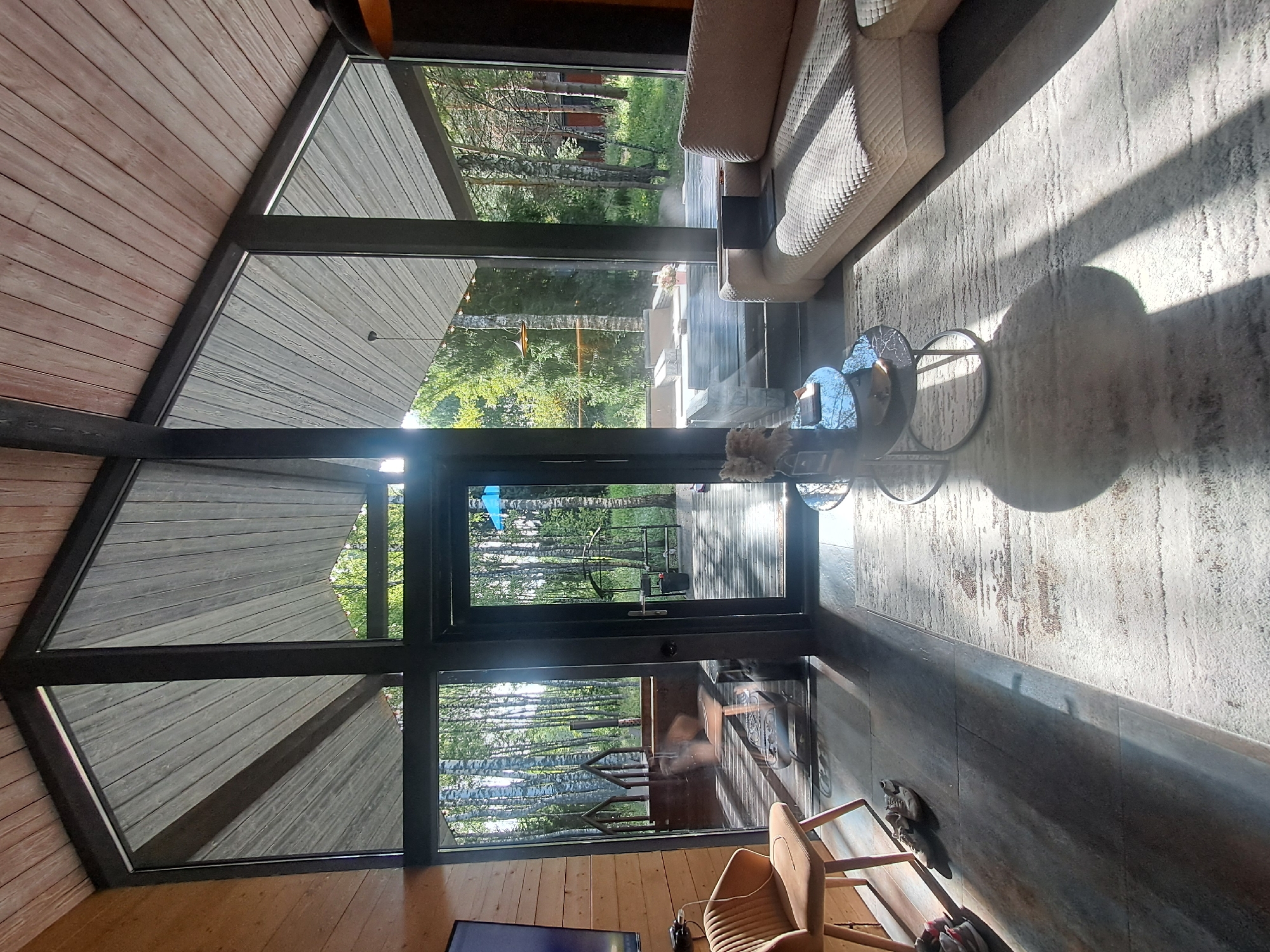 The image depicts the interior of a modern living room with a large, angled window offering a view of a wooded outdoor area. The room features a light-colored sectional sofa with a laptop on it, a small side table with a vase, and a single brown accent chair. The floor is a dark tile with a patterned rug in the foreground. Sunlight streams through the window, casting long shadows across the room. The exterior view shows numerous trees, a deck, and what appears to be a hot tub or plunge pool. The architecture of the room includes a wooden sloped ceiling. The location is identified as Krasnyi Ogorok, Russia.