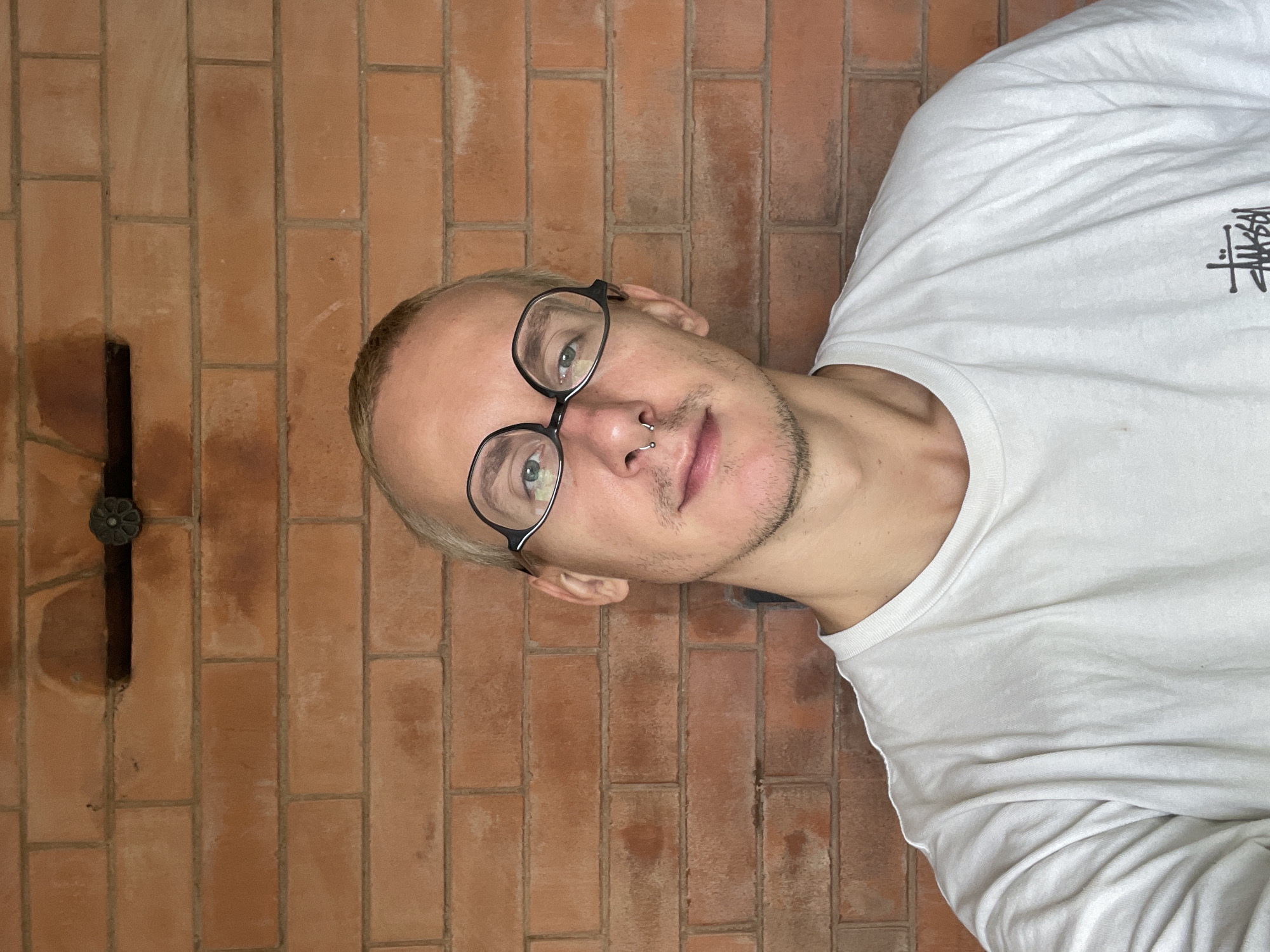 A man with short blonde hair and black-rimmed glasses poses against a brick wall. He wears a white long-sleeved t-shirt with the "Stussy" logo. A silver ring is visible in his left nostril. The brick wall has a vertical black beam with a decorative metal flower-shaped fixture. The location is unidentifiable as Sestroretsk based solely on the image content.