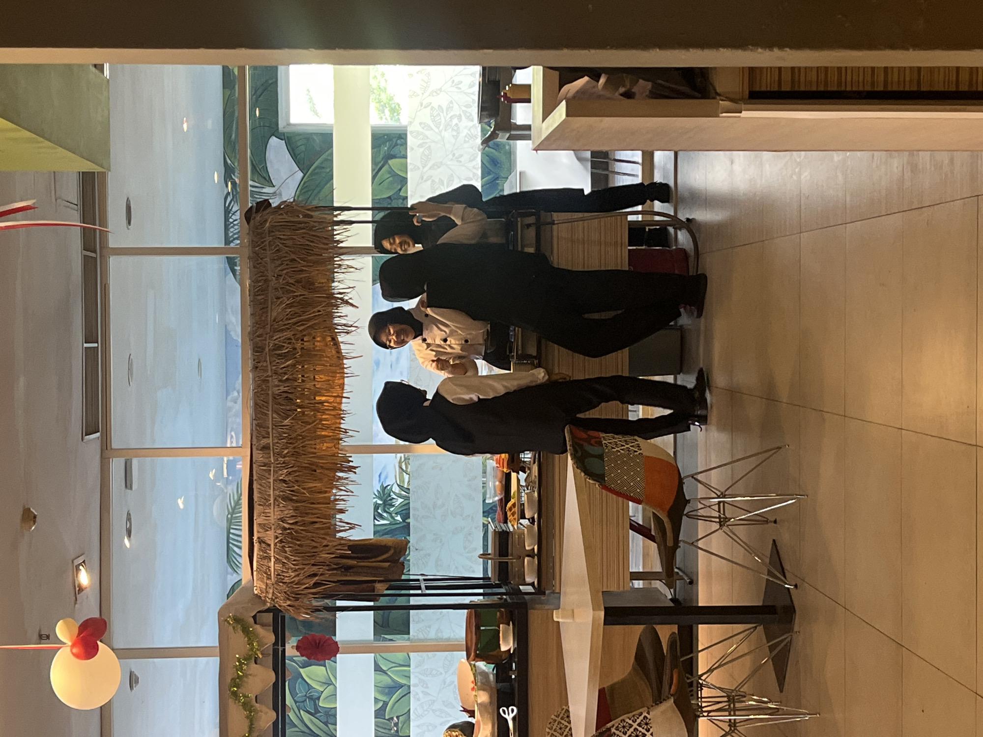 This image shows a scene at what appears to be a buffet or catering setup, possibly within a hotel or restaurant, with tropical-themed decor. The foreground features a dining table with chairs, one of which has a patchwork fabric seat. Behind the table is a buffet station with a thatched roof structure. Several individuals, seemingly staff members, are visible around the buffet. One person in a light-colored chef's coat and a black hijab is smiling and gesturing with their thumb up. Another person in a similar uniform and hijab is laughing. A third person in a black vest and hijab stands with their back to the camera. A fourth individual, also wearing a hijab, is partially visible. The background reveals large windows with a mural of tropical foliage. The location is identified as Gapurosukolilo, Indonesia.