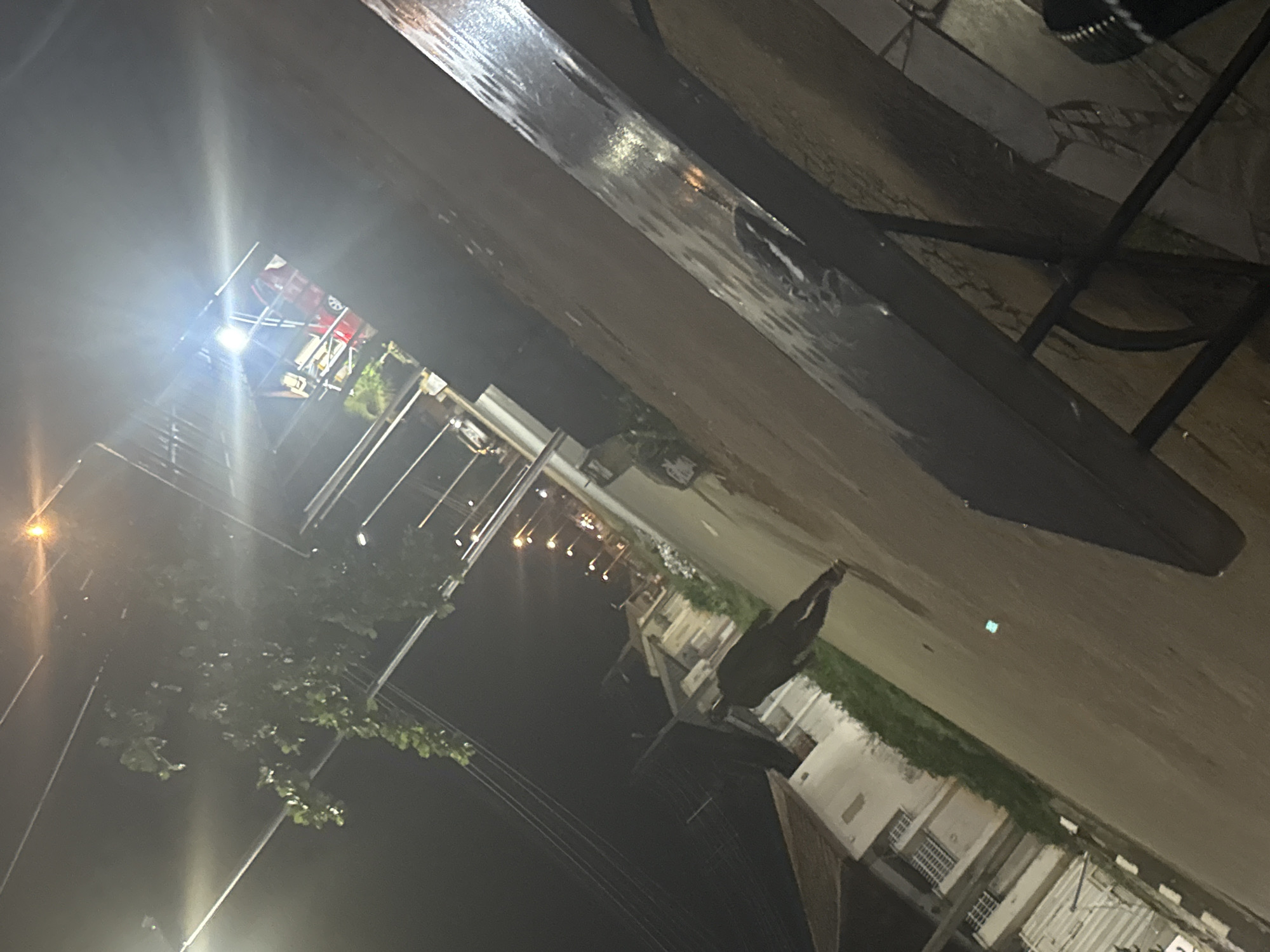 The image captures a nighttime street scene in Abuja, Nigeria. A solitary figure, dark-clothed and partially obscured, walks away from the camera down a wet, asphalt road. To the left of the road, a building with multiple windows and a tiled roof is visible. A lush green shrubbery borders the road near the building. Overhead, streetlights cast a yellowish glow, illuminating the scene. In the distance, several more streetlights are visible along the road, suggesting a developed urban area. The overall impression is of a quiet, residential street at night, with evidence of recent rainfall contributing to the wet surfaces.