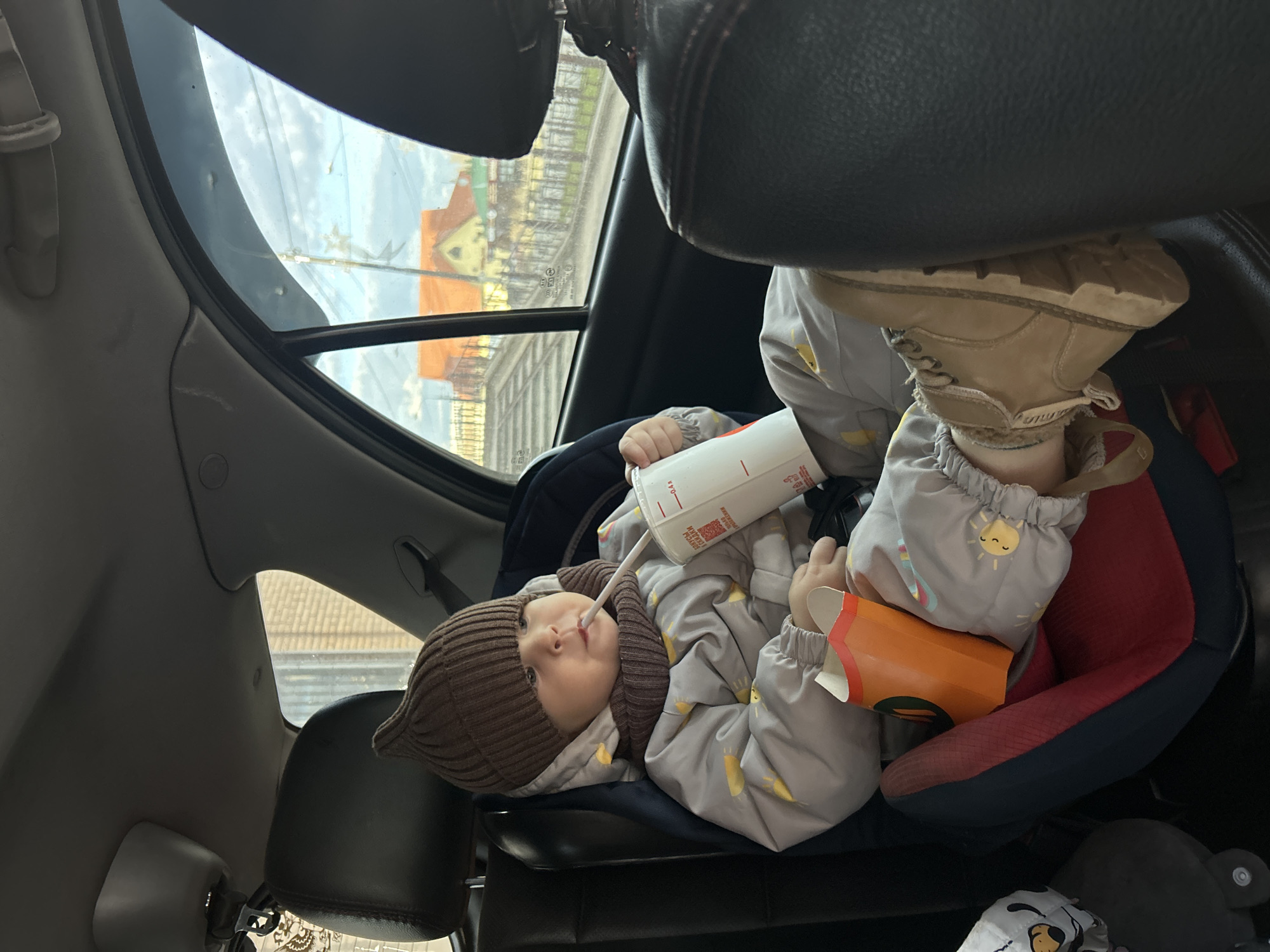 A baby wearing a brown knit hat and a gray jacket with a sun pattern is sitting in a red and blue car seat inside a vehicle. The baby is holding a white and red paper cup with a straw and an orange french fry container. The baby is holding the straw to their mouth. A brown boot is resting on the car seat. Outside the car window are buildings in the city of Klin, Russia.