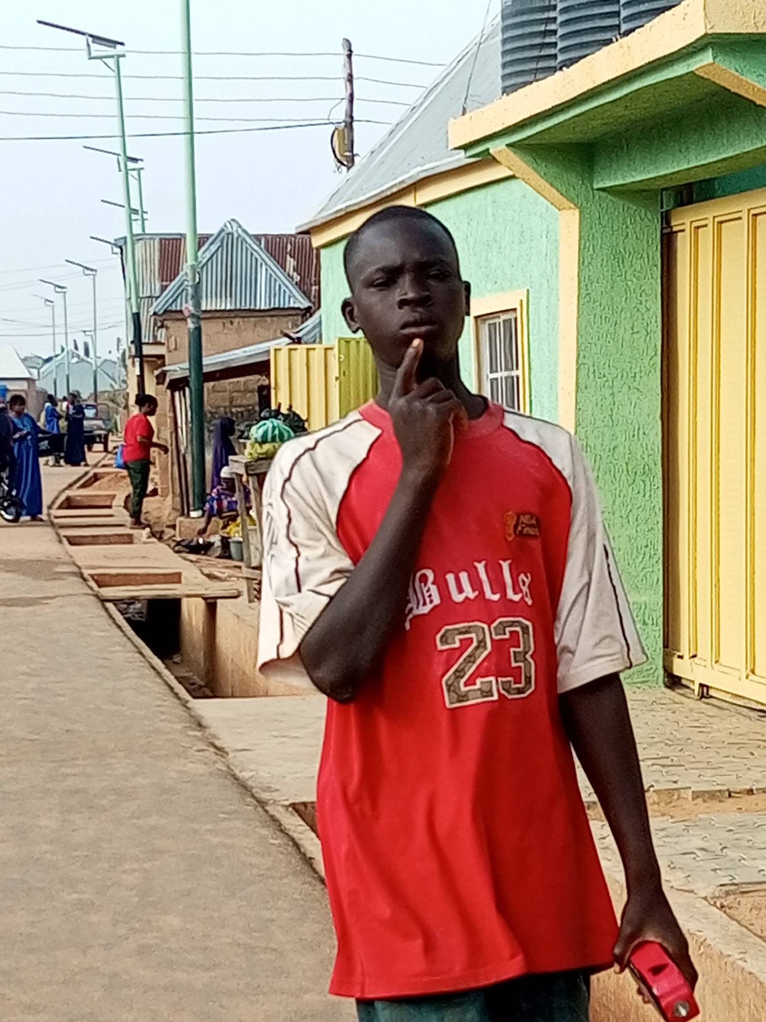 This is a medium shot, eye-level, outdoor photograph taken in Tempure, Nigeria. The focal point is a young Black male standing on a paved street, looking directly at the camera. He has dark skin and is wearing a red t-shirt with white sleeves, featuring the number "23" and "Bull" printed on the front. His left hand is raised to his chin, and his right hand is holding a red electronic device.
The background shows a street scene with buildings of various colors and architectural styles. On the left, there are several lampposts. Further down the street, people are visible, some in traditional attire. A drainage ditch runs along the side of the street. The sky is overcast.