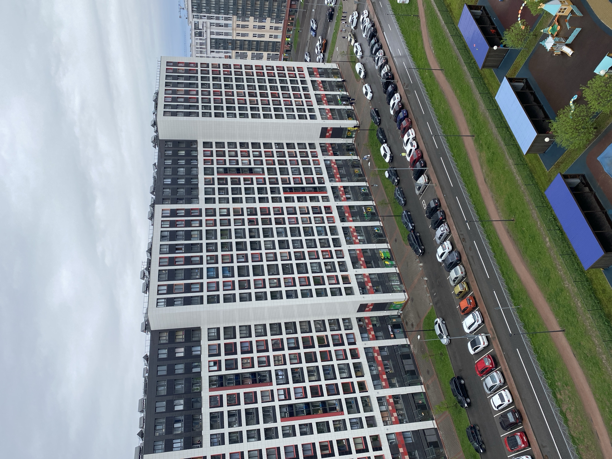The image depicts an aerial view of a residential area in an unknown city, presumed to be in Russia. The focal point is a tall, multi-story apartment building with a modern design, characterized by a grid-like facade of white, black, and red window frames. Adjacent to the building is a road lined with parked cars of various colors and models. A narrow strip of green grass separates the road from a playground area featuring blue structures and play equipment. The sky is overcast with a layer of grey clouds. No people are clearly visible.