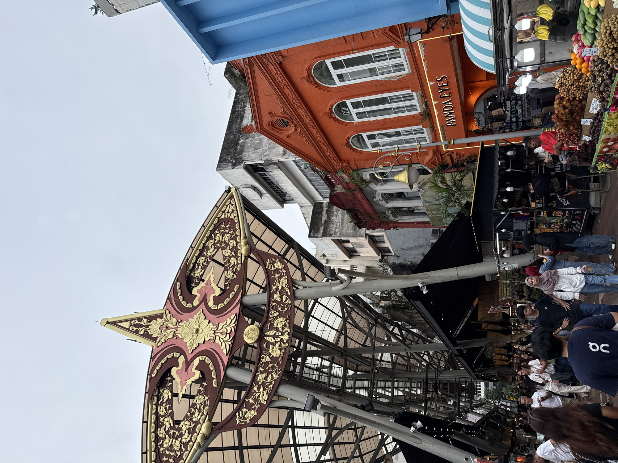 The image depicts a street scene in Kuala Lumpur, Malaysia, known for its architectural fusion and vibrant street life. An elaborate ornamental structure, possibly part of a market or pedestrian area, features a brown and gold floral design under a translucent roof. Buildings frame the view, including an orange building with white windows labeled "PANDA EYES" and a blue structure. A fruit stand displays various tropical fruits. A crowd of people strolls through the street, some interacting with vendors or each other. The setting suggests a lively, possibly touristic area in the city.