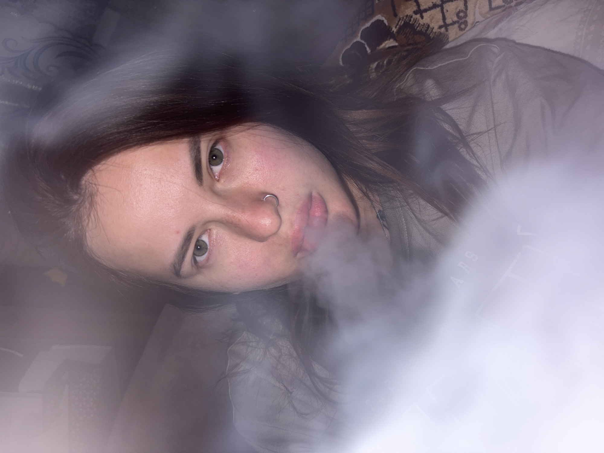 A person with long brown hair and a nose ring is pictured against a backdrop of a bed with patterned sheets. The person is exhaling smoke towards the camera. The location is Unknown, Unknown.