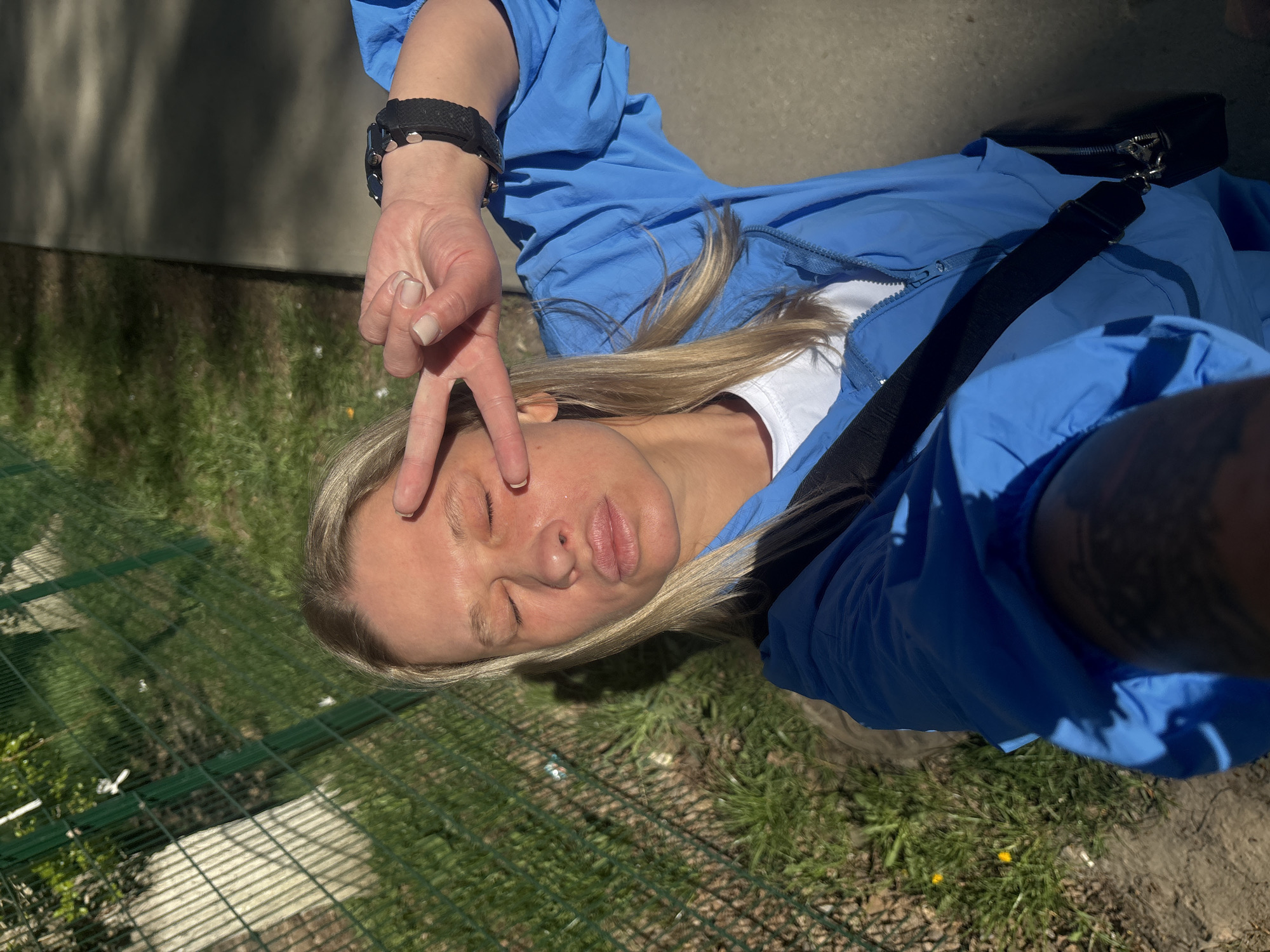 A selfie captures a fair-skinned woman outdoors, angled so she appears prone. She is dressed in a blue jacket over a white top. Her blonde hair is styled loosely. Her right hand is raised, displaying a "peace" sign near her closed eyes. The woman wears a black watch or bracelet on her left wrist. A black strap of some kind is visible across her body. The background shows grass and a section of green metal fence. The location is the city of Perm, Russia.