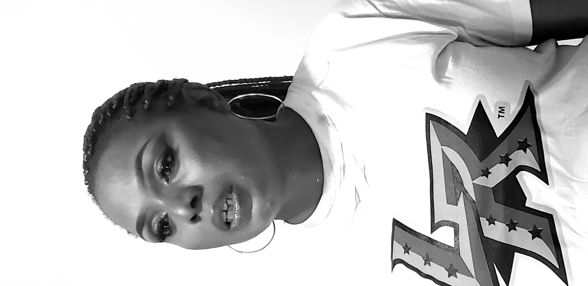 A black and white medium shot shows a Black woman wearing a white t-shirt with a graphic design on it. She has braided hair and is wearing large hoop earrings. She is looking to her left with her mouth slightly open, as if speaking. The background is a plain white wall.