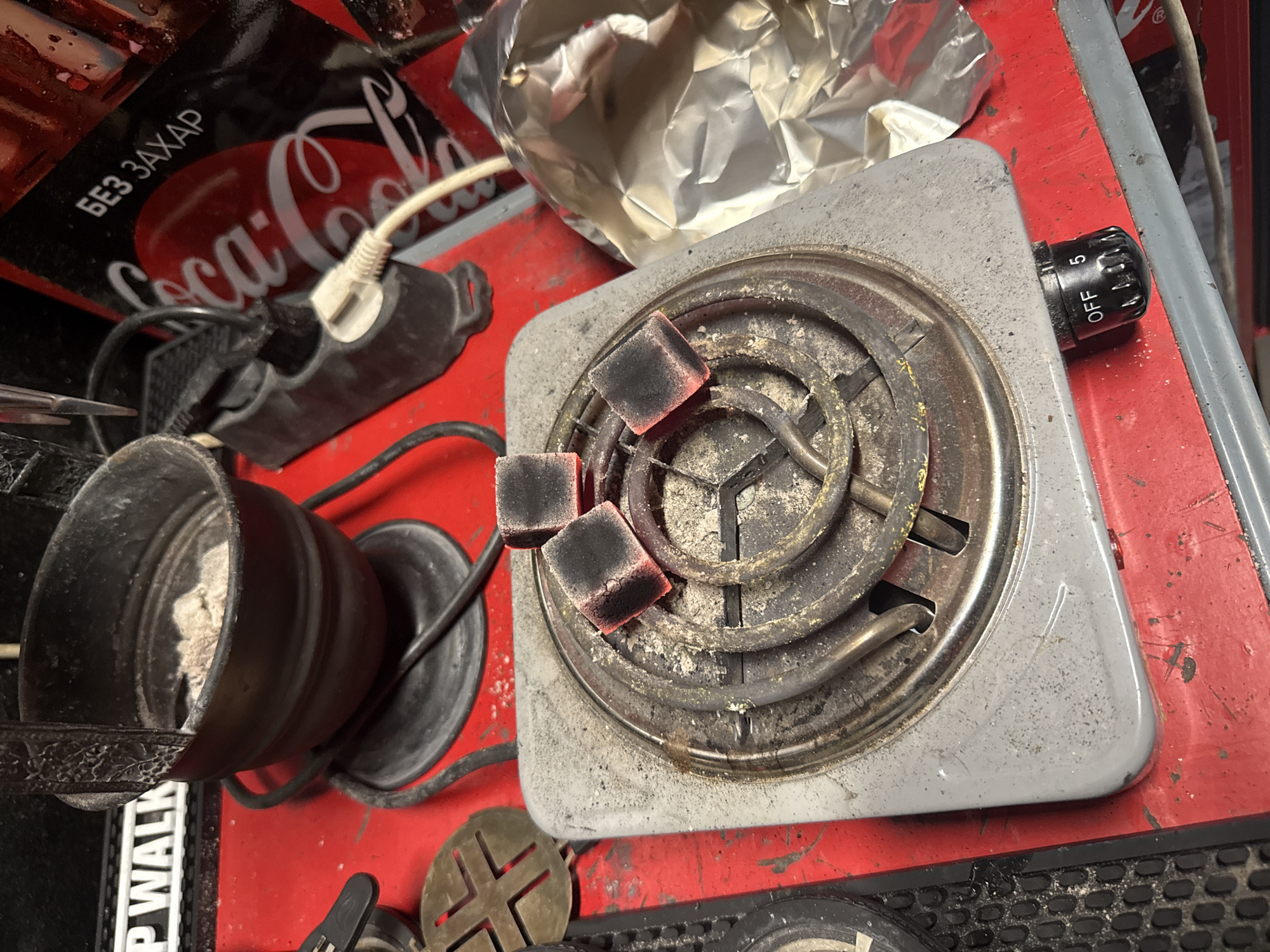The image captures a close-up, overhead view of items resting on a red, Coca-Cola branded surface, likely a tray or cooler. The central subject is a small, gray electric hot plate with visible heating coils. Three square charcoal briquettes are placed on the coils, two of which are glowing red, indicating they are actively heating. Ash residue is present on and around the burner.
To the left of the hot plate, a black metal cup or container, partially filled with ash, stands upright. An electrical cord extends from an outlet in the background towards an electrical plug, which is connected to a black object. Another black cord runs from the hot plate.
In the upper left background, part of a black Coca-Cola product is visible, bearing the white text "Coca-Cola" and "БЕЗ ЗАХАР," which is Cyrillic for "NO SUGAR." A crumpled piece of aluminum foil is situated behind the hot plate, slightly to the left. The hot plate's control knob, labeled "OFF" and "5," is on the right side. Below the hot plate, a dark metallic object with a cross-shaped pattern can be seen, along with a black textured strip partially showing "P WALK." The overall setting appears to be an indoor space, and the items suggest preparations for a hookah or similar activity where charcoal heating is required. The surfaces show some wear and tear, with dust and scuffs visible on the hot plate and the red tray.