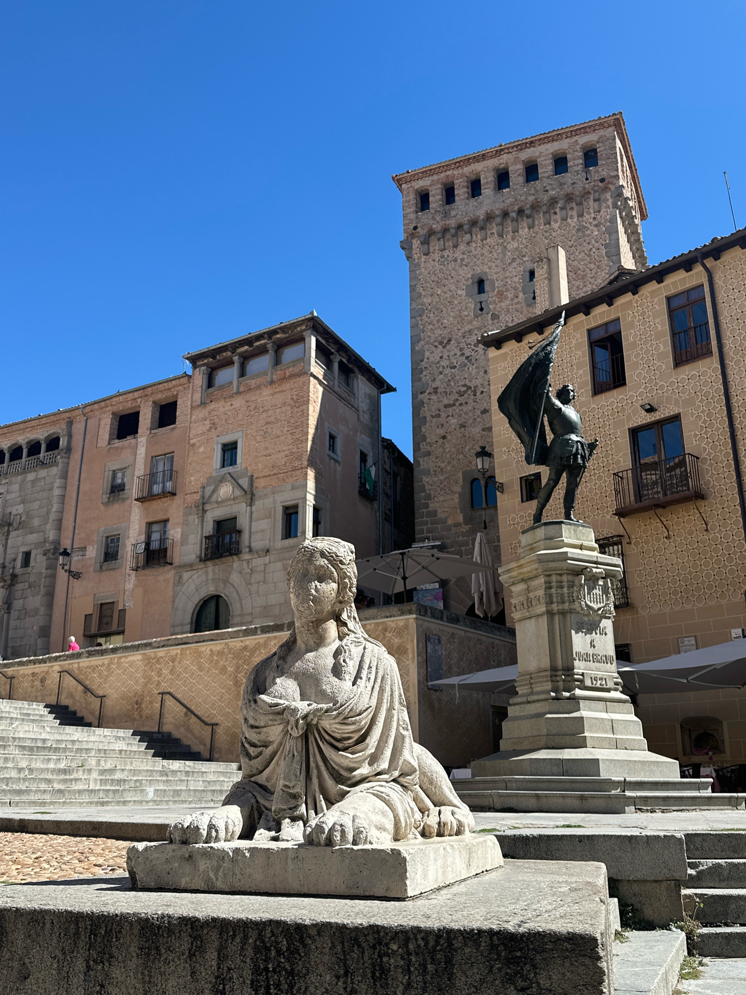This is a photograph of a stone sphinx statue in the foreground, with a monument topped by a statue of a man holding a flag in the mid-ground. Behind the monument are historic buildings, one of which is a tall stone tower. The sky is clear and blue. The scene is outdoors, likely in a public square or plaza. The sphinx is a light-colored stone and is depicted in a reclining position with paws extended. The monument is also stone, with an inscription on its base. The statue on top of the monument is of a man in historical attire, holding a dark flag aloft. The buildings have a mix of stone and plaster facades, with windows and balconies. The overall impression is of an urban historical site. This scene is located in Plaza del Acueducto in Segovia, Spain, not La Lastrilla, Spain.