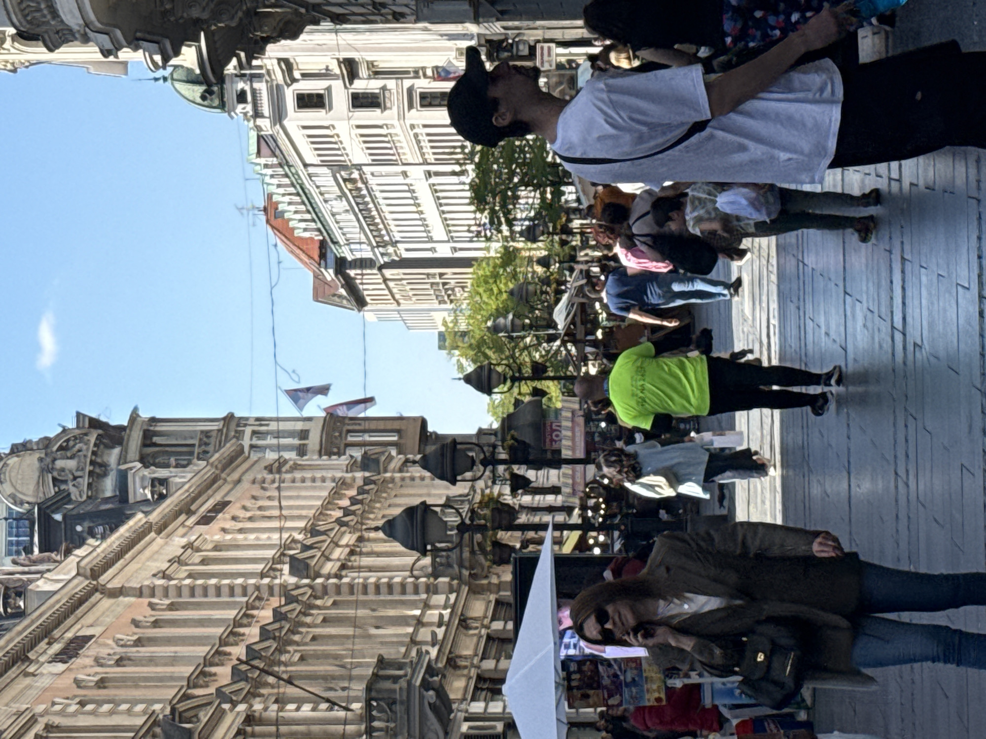 The image captures a vibrant, sunlit street scene in a European city, confirmed by context as Belgrade, Serbia. Numerous people populate a wide, paved pedestrian area flanked by ornate, multi-story buildings.
**Subjects:** The foreground features a woman with long dark hair, wearing sunglasses, a dark jacket, and blue jeans, looking down at a smartphone while holding a black shoulder bag. Adjacent to her, a large white umbrella or awning is partially visible. Further into the scene, a man in a bright lime green t-shirt and black pants walks past street lamps. In the upper right, a man in a white t-shirt and black baseball cap looks upward, with a classical-style building facade behind him. Many other pedestrians, whose faces are mostly obscured, fill the background, indicating a busy thoroughfare. The buildings exhibit classical architecture with intricate details, columns, cornices, and balconies. One building on the left side features a green dome.
**Activity or event:** The scene depicts typical daily life in a city center. People are walking, standing, and engaging in casual activities, suggesting a bustling urban environment, possibly a shopping street or a promenade.
**Notable details:** The time of day is bright and sunny, indicated by the clear blue sky with minimal clouds and strong lighting on the buildings and people. The weather appears pleasant. Two flags, resembling Serbian flags with red, blue, and white stripes, are visible hanging from a building on the left, serving as a location cue. Black, multi-armed street lamps are intermittently placed along the street. Some greenery from trees adds a touch of nature amidst the urban landscape.
**Visible text:** Faint Cyrillic text is discernible on a sign in the middle ground, where the word "БОЛ" (BOL) is partially legible, likely part of a larger sign.