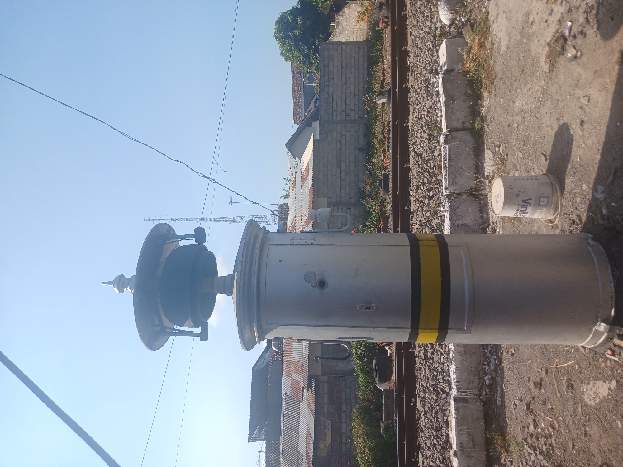 The image depicts an outdoor scene in Banyuwangi, Indonesia, likely near a railway line. In the foreground, a tall, silver cylindrical object with a decorative top stands prominently. This object has two horizontal bands, one yellow and one black, around its middle section, and what appears to be a door or access panel on its side. Beside it, a white plastic bucket with some markings and a coiled rope is resting on the dusty ground.
In the midground, railway tracks are visible, laid on a bed of gravel. Beyond the tracks, a variety of buildings are present, characterized by corrugated metal roofs and brick walls. Some low-lying vegetation and a partially visible discarded tire are also scattered in this area. In the background, a clear blue sky dominates, with a distant communication tower and power lines stretching across the frame. The overall impression is one of a functional, perhaps industrial or transportation-related, area on a sunny day.