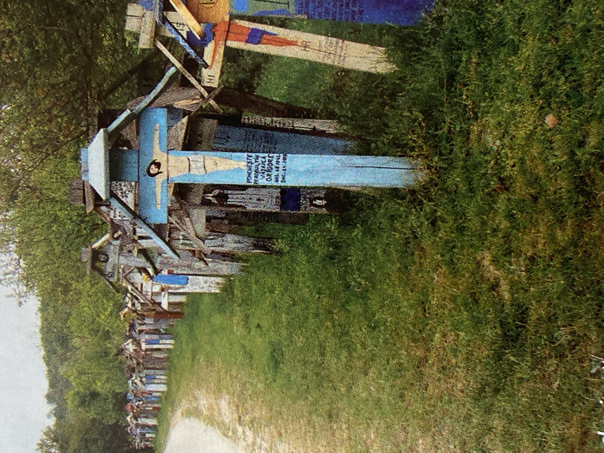 The image depicts a roadside lined with numerous wooden crosses, creating a cemetery-like atmosphere. The crosses vary in size and design, with some featuring painted imagery of Jesus Christ. Many of the crosses are adorned with inscribed text, likely names and dates. The scene is set in an outdoor environment with a dirt path to the left and lush green grass and trees in the background. The lighting suggests it is daytime, with a slight overcast sky.