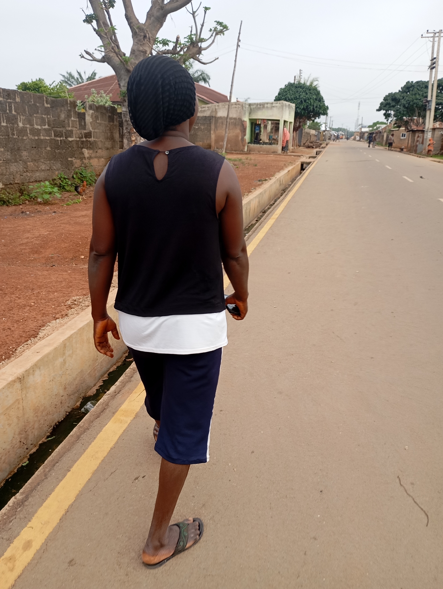 The image depicts a rear view of a person walking away from the camera down a paved road in Shendam, Nigeria. The individual is wearing a black sleeveless top, a white undergarment, dark blue shorts with white trim, and dark flip-flops. They are holding a dark-colored object, possibly a phone, in their right hand. The road has a yellow line marking the edge of the lane and a concrete gutter with some debris runs alongside the road to the left. The environment includes brick walls, buildings, trees, and utility poles, suggesting a semi-urban or rural setting. The sky is overcast. In the background, other people are visible walking along the road. A chicken is present in the foreground near a brick wall.