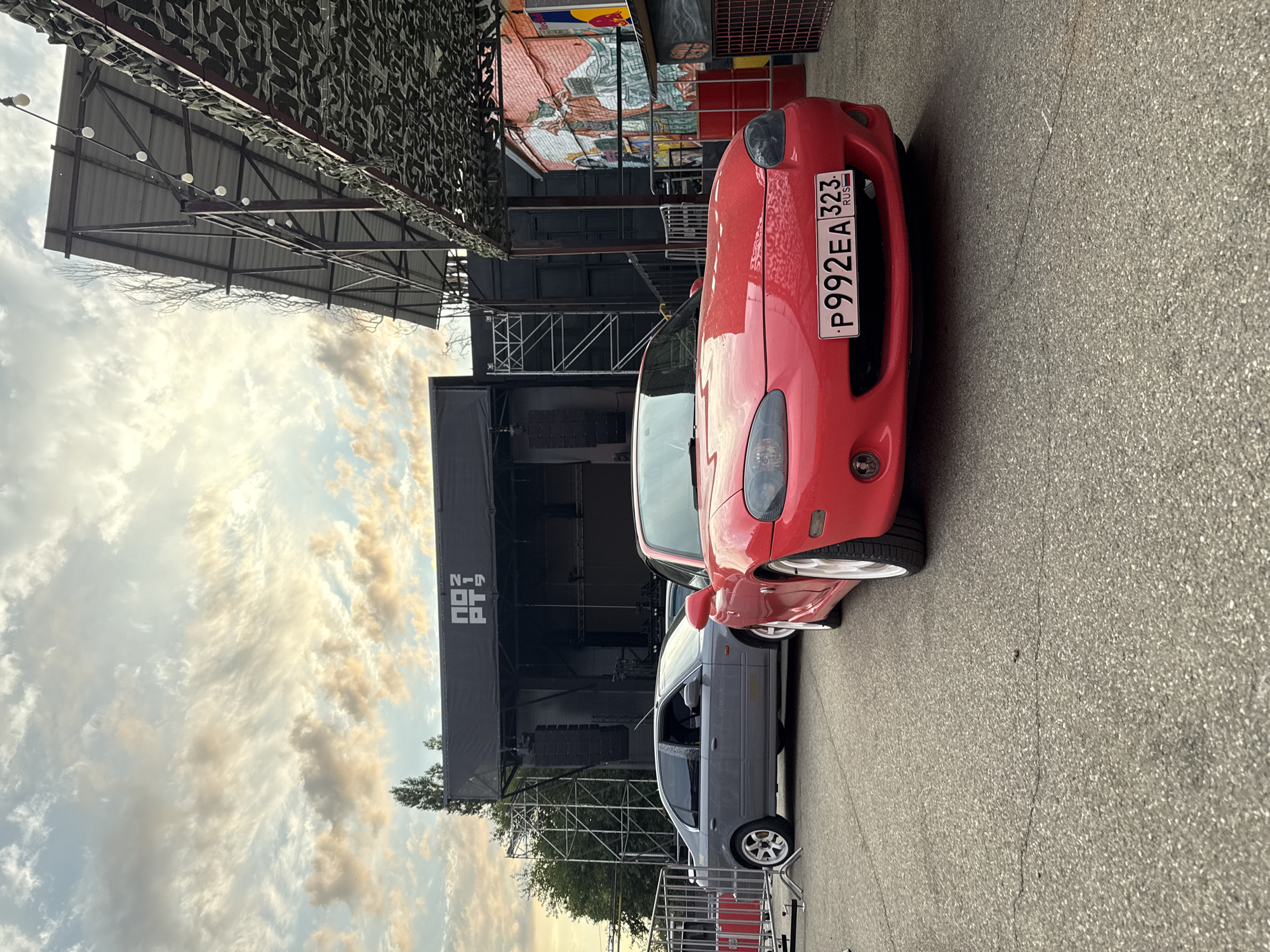 This low-angle, eye-level, outdoor photograph depicts two cars parked on asphalt in front of a black stage structure. The car in the foreground is a red Mazda Miata with a license plate reading "P992EA 323 RUS". To its left, a gray sedan, possibly a Honda Civic, is parked. Behind the cars, a large black stage is set up with scaffolding and sound equipment. The sky above is partly cloudy with warm tones, suggesting either sunrise or sunset. The scene is set in an urban or event location. The overall impression is that of an outdoor event or concert setup with vehicles present. The specific location in Krasnodar, Russia is not discernible from the image itself.