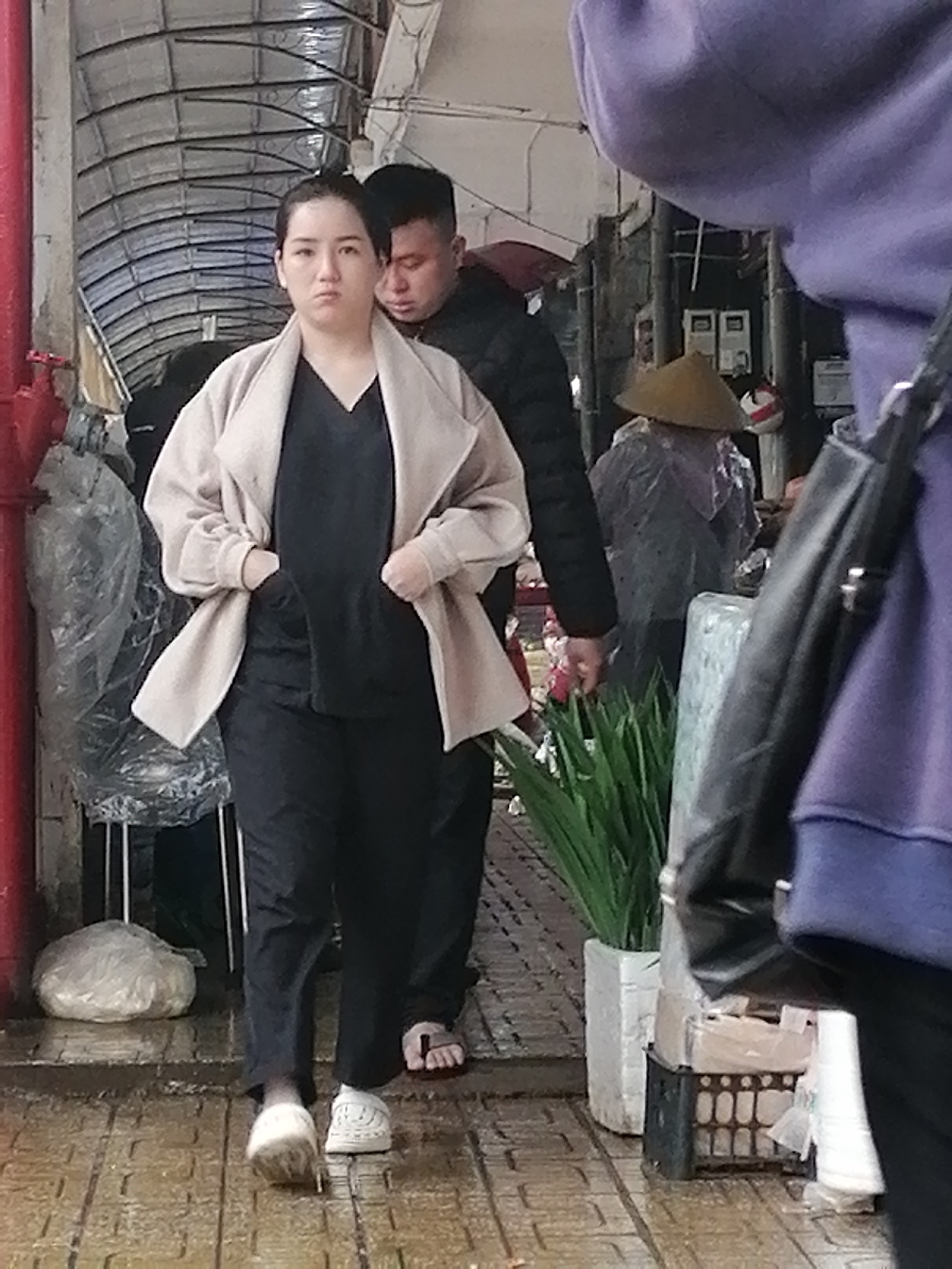 The image captures a scene at a market in Da Lat, Vietnam. In the foreground, a woman with her hands in the pockets of her light-colored coat and black pants walks towards the viewer. She has her hair pulled back and a neutral expression. Behind her, a man in a dark jacket is partially visible. To the right, a person wearing a purple hoodie is seen from the shoulders down, with a black bag slung over their shoulder. Further back, another individual is visible wearing a conical hat and what appears to be a plastic poncho. The environment suggests an outdoor market setting with tiled flooring, some covered areas with corrugated metal roofs, and various objects like plastic-wrapped items, potted plants, and crates.