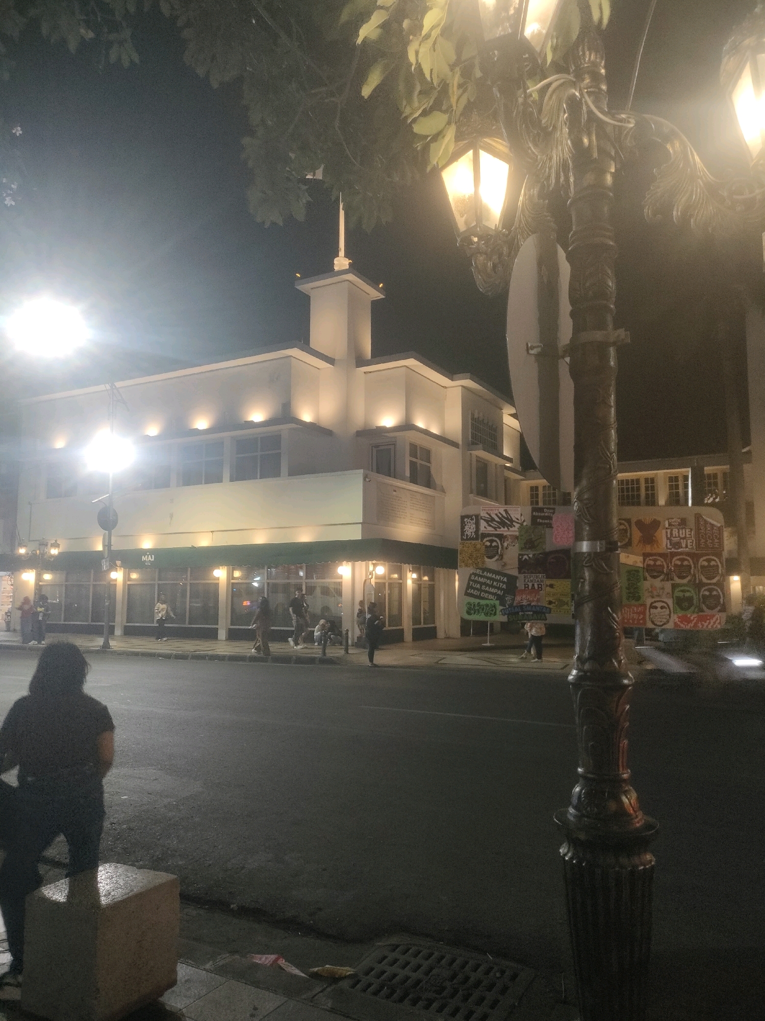 The image captures a nighttime street scene in Surabaya, Indonesia, focusing on a multi-story building with illuminated windows and decorative lighting. In the foreground, a decorative street lamp with multiple lights stands prominently on the right side of the frame. A person, silhouetted against the dim light, is partially visible on the left side, facing away from the camera and standing near a stone planter. Several other individuals are scattered along the sidewalk and on the street in front of the building. The building itself features a modern facade with distinct horizontal lines and is illuminated by spotlights. A green awning extends across the lower level of the building. Signs with colorful graphics and text are displayed on the side of the building, suggesting a commercial or cultural space. The street is paved and appears wet in places, reflecting the ambient light. Trees with foliage overhang parts of the scene, contributing to the nighttime ambiance.
