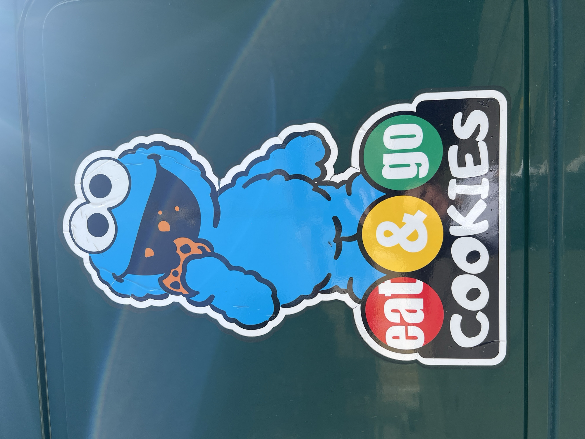 A sticker is affixed to a dark green surface. The sticker features a depiction of the Cookie Monster, a blue furry character from the children's television show Sesame Street, with large googly eyes and an orange polka-dotted bowtie. Beside the Cookie Monster is a graphic with three colored circles containing text: "eat" in red, "&" in yellow, and "go" in green, stacked vertically. To the right of the circles, "COOKIES" is written in white block letters on a black background with a white border. The sticker appears slightly wrinkled and faded. The background surface is a plain, dark green, and the image is taken from a slightly elevated angle. The sticker may be on a vehicle. The location cannot be determined based on the image alone.