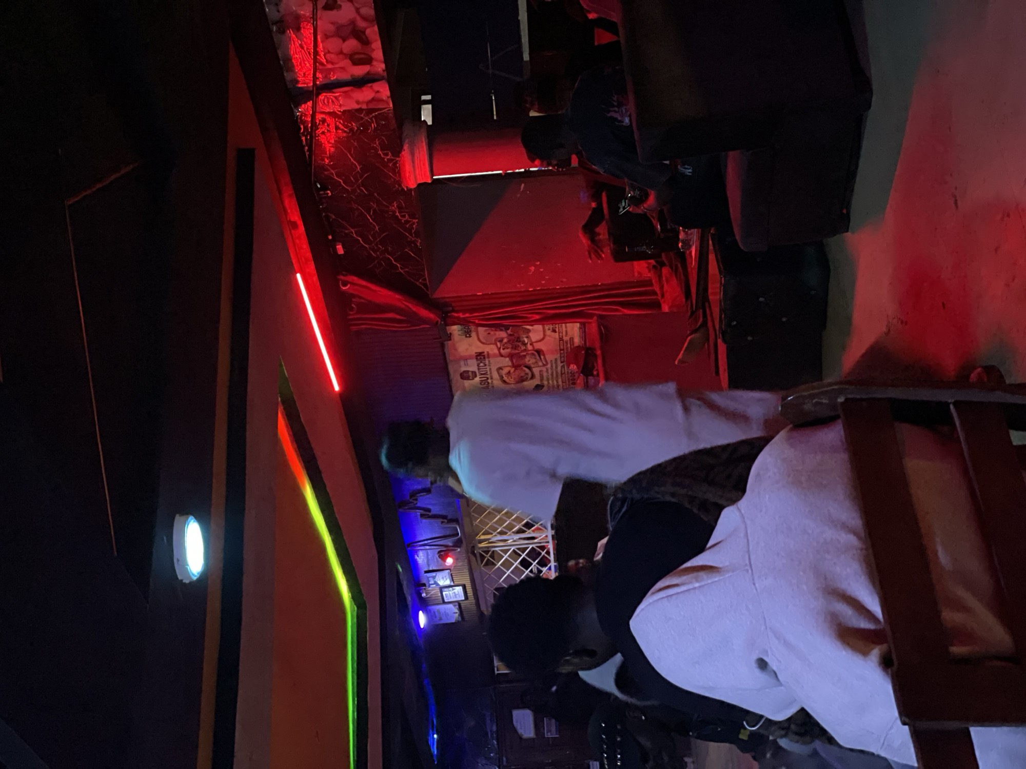 The image depicts a dimly lit, indoor scene, likely a lounge or entertainment venue, with heavy use of red and neon lighting, including red accent lighting and green neon strips. The atmosphere appears somewhat informal and possibly late-night, given the low light.
The subjects are several individuals, their faces mostly obscured or turned away from the camera, suggesting a candid or casual moment. One person in the foreground is wearing a light-colored hooded sweatshirt, with their head bowed. Behind them, another person is seen from the back, also appearing to be seated. Further back, a person in a white garment is partially visible, possibly standing or moving. The presence of multiple people suggests a social gathering.
Objects visible include furniture, such as chairs and possibly couches, and what appears to be a menu or poster mounted on the wall in the background, though the text is too blurry to read clearly. The overall setting is suggestive of a place where people gather to relax or socialize.
The image appears to have been taken at night, as indicated by the interior lighting dominating the scene. There are no clear indicators of weather conditions. The location context provided is Durimi, Nigeria. No discernible emotions are evident due to the lighting and obscured faces.