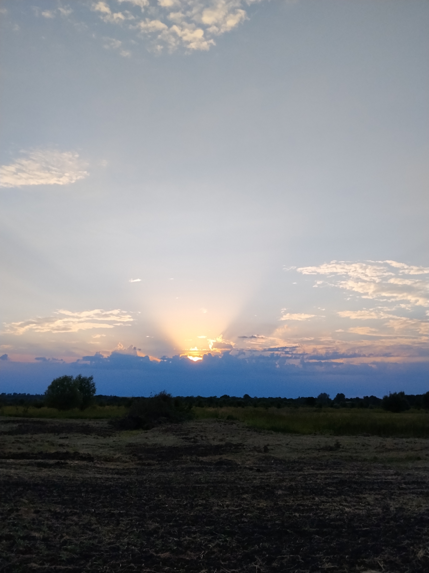 The image shows a rural landscape at sunset. The sky is a gradient of pale blue to warm orange, with scattered white clouds. The sun is partially obscured by dark clouds on the horizon, casting dramatic rays of light upwards. In the foreground, a field of dark, tilled soil is visible. Behind the field, a line of trees and bushes forms the distant horizon. The overall mood of the image is serene and peaceful, characteristic of a sunset. The time of day is clearly sunset, with the low angle of the sun and the warm colors in the sky. There are no people or text visible in the image. The location appears to be a rural area with agricultural land.