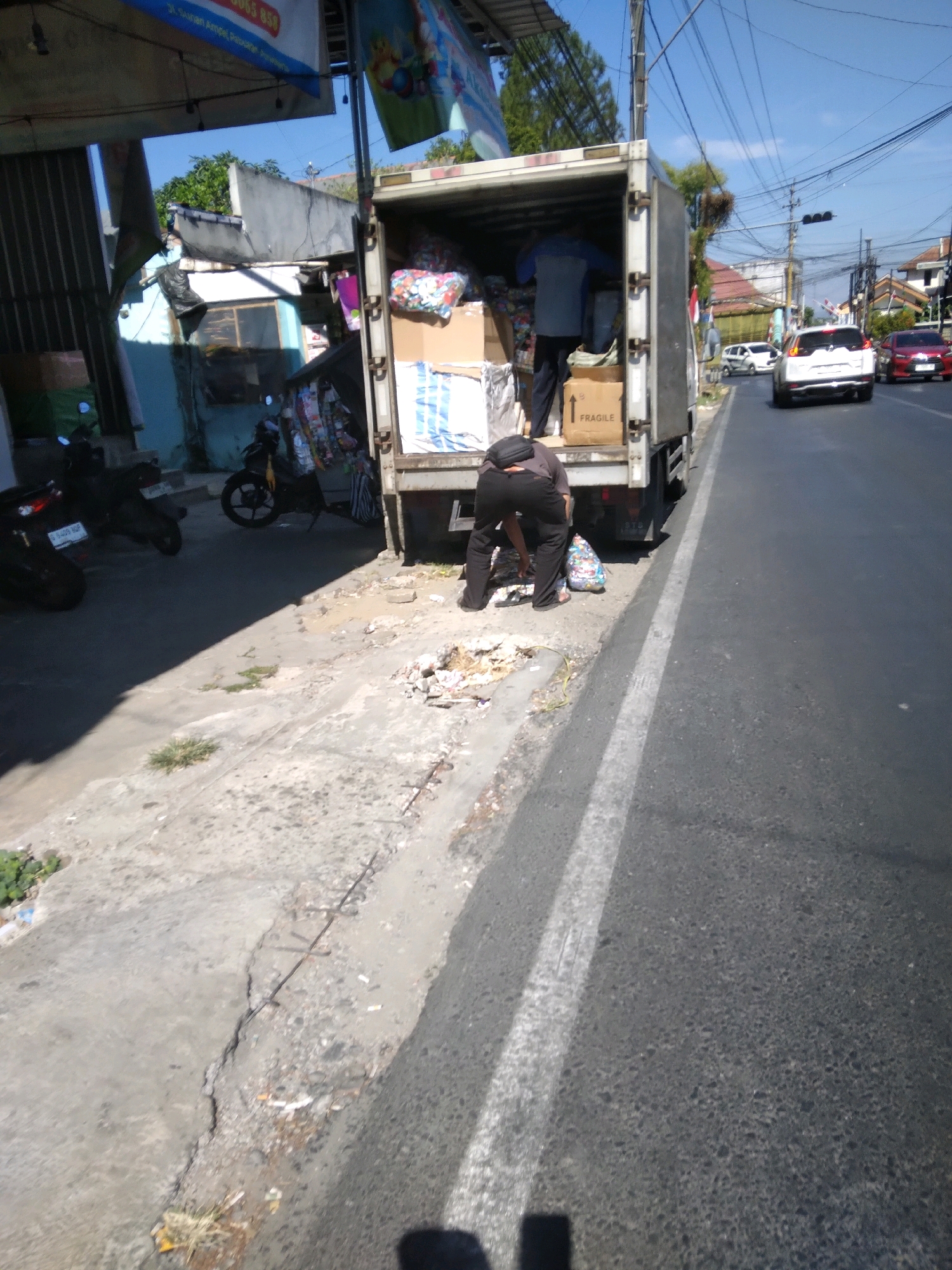 In Kedungmalang, Indonesia, a man wearing black pants and a dark t-shirt is bent over next to the open rear of a white delivery truck. He is picking up a colorful bag from the ground. Inside the truck, several boxes are visible, one of which is labeled "FRAGILE" with an upward-pointing arrow. Another man, wearing a blue and grey shirt and dark pants, is also inside the truck, appearing to organize or load items. The truck is parked on the side of a road, with two motorcycles parked on the left side of the scene. In the background, houses, power lines, and other vehicles are visible, suggesting an urban or semi-urban environment. The sky is clear and blue, indicating a sunny day. The ground is a mix of concrete and dirt, with some debris scattered around.