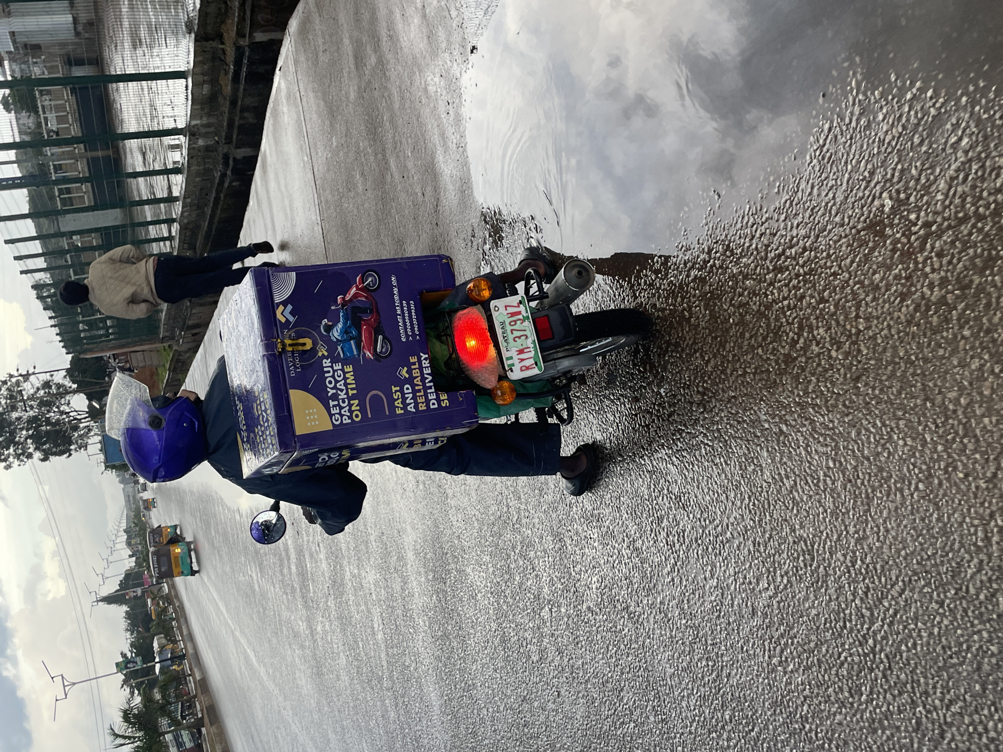 In Barakin Lerawi, Nigeria, on a wet asphalt road, a person wearing a dark blue uniform and a purple helmet is seated on a motorcycle. The motorcycle has a large purple storage box on the back, branded with "DAVEBUN LOGISTICS" and text stating "GET YOUR PACKAGE ON TIME" and "FAST AND RELIABLE DELIVERY". The license plate of the motorcycle reads "RYM 379NZ PLATEAU". The road is wet, with large puddles reflecting the overcast sky. In the background, a man in a light brown jacket and dark pants walks away from the camera. Other vehicles, including a yellow auto-rickshaw, are visible further down the road. The environment appears to be an urban or semi-urban setting with some greenery and structures along the roadside.