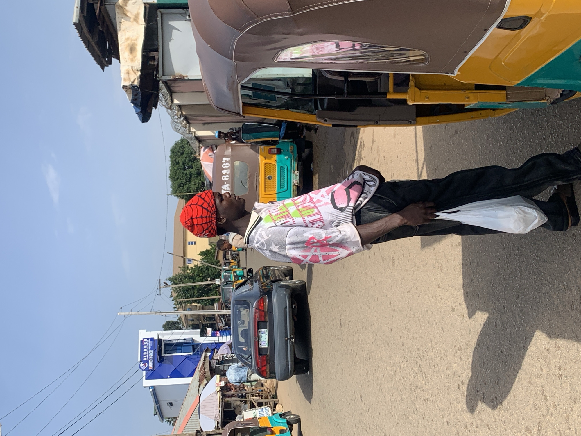 In Jos, Nigeria, a man wearing a red cap with a spider web pattern stands on a dusty street next to a yellow and brown auto-rickshaw. He is wearing a white t-shirt with a pink and black graphic print and dark pants. He holds a white plastic bag in his right hand. The street is lined with buildings and stalls, and other vehicles, including a dark sedan and other auto-rickshaws, are visible in the background. The sky is clear and blue. The overall scene depicts a bustling street in a Nigerian city.