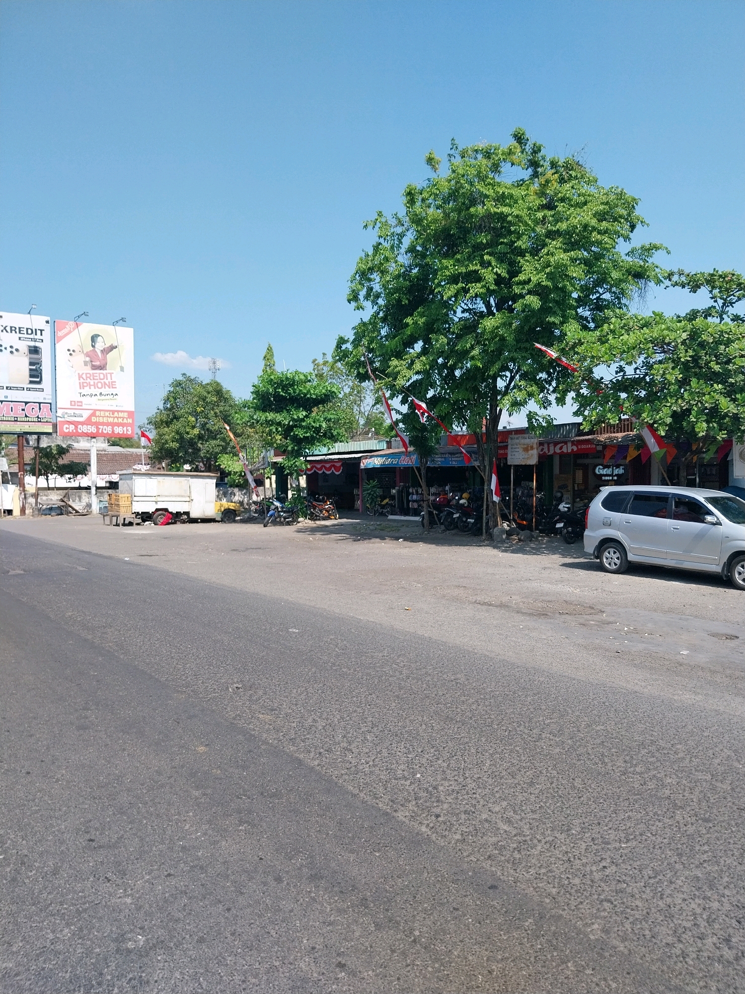 The image depicts a street scene in Gondang Kulon, Indonesia, under a clear blue sky. On the left side of the road, a large billboard advertises "KREDIT IPHONE Tanpa Bunga" (iPhone Credit Without Interest) with a contact number. Adjacent to the billboard is a white truck with a cargo box. Further down the street, several small shops with red and white Indonesian flags are visible. These shops appear to be retail establishments selling various goods, with motorcycles parked outside and in front of them. A silver minivan is parked on the right side of the road in front of one of the shops. The road surface is asphalt, with some patches of discoloration and scattered debris. Lush green trees line the area behind the shops. The overall scene suggests a typical roadside commercial area in a town or city in Indonesia.