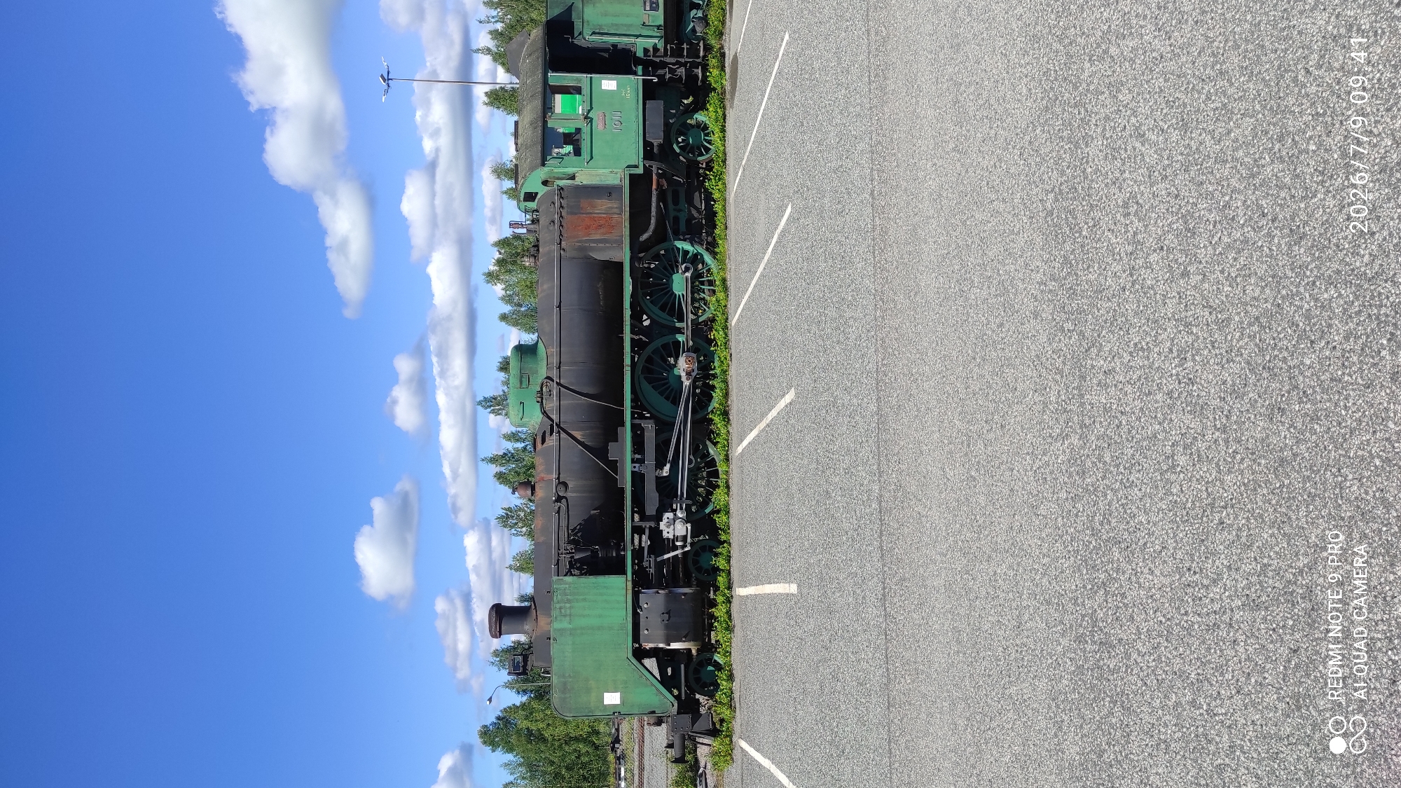 An old, green and black steam locomotive sits on tracks in a paved area in Otanmäki, Finland. The locomotive is partially covered in green paint, with areas of rust visible on the boiler. It has large, spoked wheels and a visible cabin. The background consists of a line of trees under a blue sky with scattered white clouds. The foreground is a paved surface marked with white lines, resembling a parking lot. There are no people visible in the image.