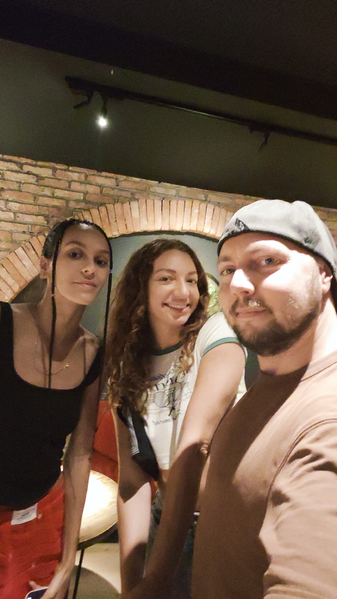 This is a selfie of three people, two women and one man, taken indoors. The background features a brick archway and a dark ceiling with some lighting fixtures. The man is wearing a baseball cap and a brown t-shirt. One woman has braided hair and is wearing a black tank top and red pants. The other woman has curly brown hair and is wearing a white t-shirt with a green trim. All three are smiling. The lighting suggests it is likely evening or indoors with artificial lighting. There is no visible text in the image.