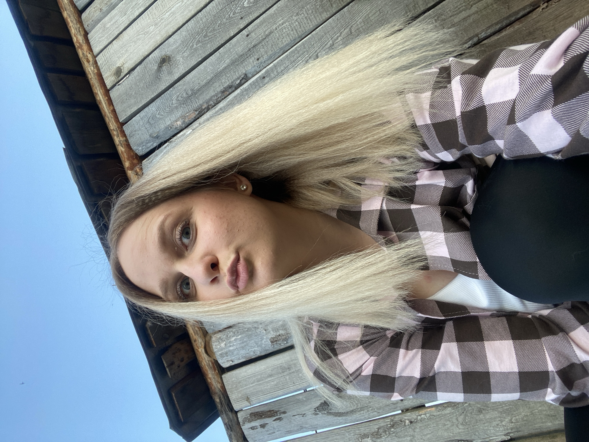 The image is a low-angle portrait of a young woman, presented in a tilted orientation, rotated approximately 90 degrees clockwise. She has long, straight, blonde hair that spreads across a weathered wooden surface behind her. Her light-colored eyes are directed towards the camera, and her expression is neutral to slightly pensive, with her lips softly pursed. She is wearing a plaid shirt with a pattern of brown and light pink squares, layered over a white top. A dark, possibly black, garment is visible below her plaid shirt on the right side of the frame.
The background consists of a rustic wooden structure made of horizontal planks, which appear aged and textured. A rusted, dark brown metal bar or pipe runs diagonally across the upper portion of the wooden background, suggesting it is part of an older building or fence. To the left of the wooden structure, a clear, bright blue sky is visible, with a small, distant airplane subtly silhouetted in the upper left corner of the sky. The natural lighting suggests it is daytime with good weather. No text is visible in the image. The scene is outdoors, consistent with a rural or semi-rural setting in Kauchuk Stantsiya, Russia.