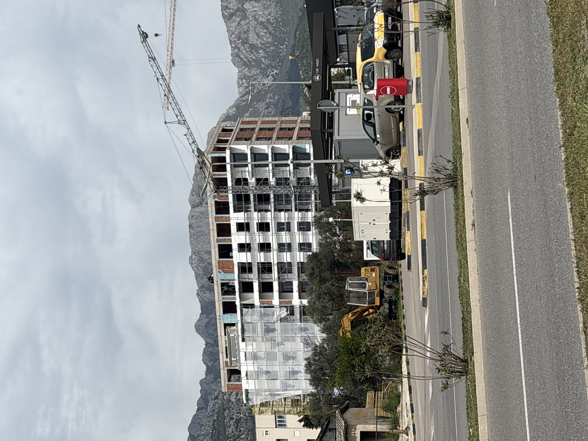 A mid-level, slightly oblique outdoor shot depicts an unfinished, multi-story building under construction in the city of Bar, Montenegro. The building’s structure is visible with exposed brickwork on the upper floors, and scaffolding covers the lower sections. A construction crane looms over the structure. In the foreground, there's a road with a white line divider, bordered by a yellow and black curb. Vehicles, including cars and possibly a car wash structure, are parked along the road. A mountainous landscape provides the backdrop behind the building. The sky is overcast, suggesting a cloudy day. Additional construction equipment, including an excavator, is parked near the building. Some vegetation is also visible near the road and the construction site.