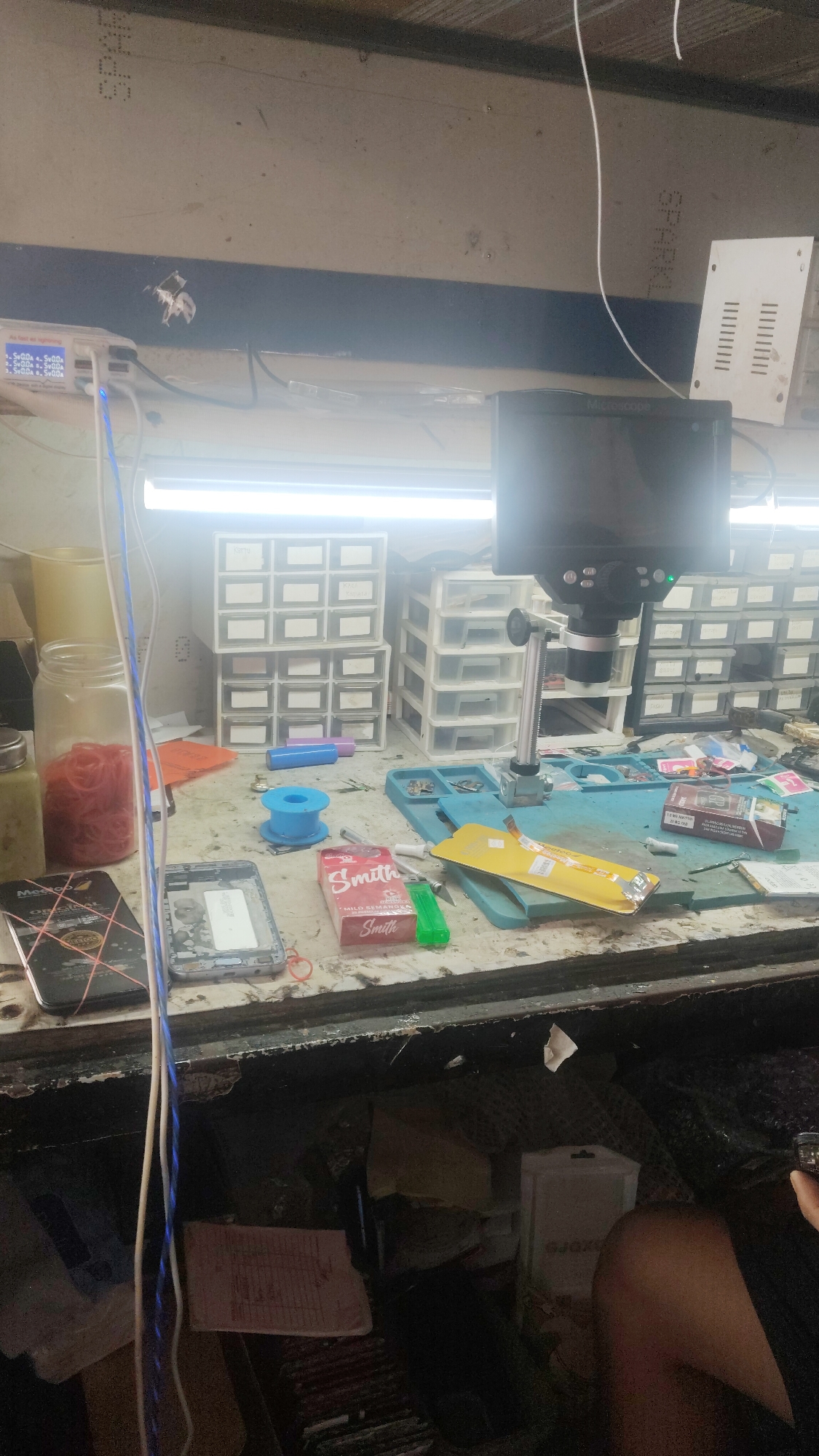 The image depicts a cluttered workbench, likely in a repair shop, in Bandar Lampung, Indonesia. The workbench is covered with various electronic components, tools, and personal items. In the foreground, a disassembled mobile phone is visible, along with a pack of "Smith" cigarettes and a green lighter. Behind these items, there are stacks of plastic organizer drawers containing small parts, and a microscope is set up for detailed work. Wires and cables are strewn across the surface, suggesting ongoing electronic repair activities. The background shows shelves filled with more components and equipment, typical of a service center or workshop environment. The overall scene is one of organized chaos, indicative of a busy workspace.