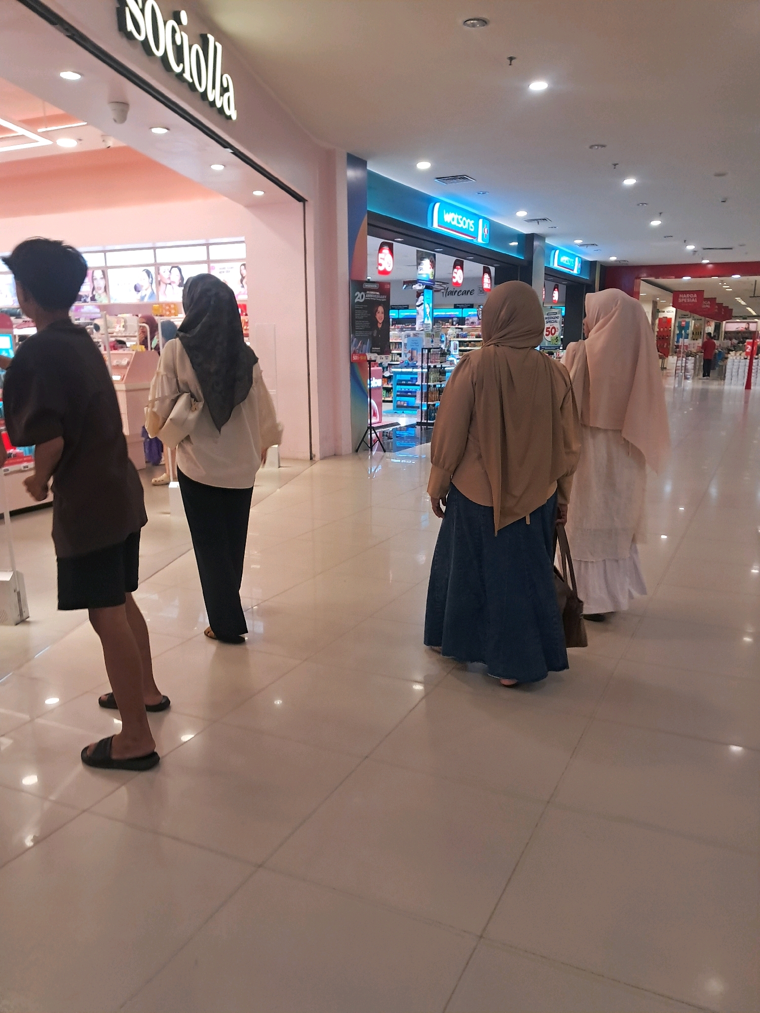 The image captures a scene inside a brightly lit indoor shopping mall, likely in Singkawang, Indonesia, judging by the "SOCIolla" store sign which is a popular beauty retail chain in Indonesia. The perspective is from behind a group of four individuals walking away from the camera.
On the left, a person wearing a dark t-shirt and black shorts, with dark hair, walks with their back to the viewer, their feet clad in black slides. To their right, a person wearing a cream-colored long-sleeved blouse and black pants, with a black hijab and a light-colored handbag, walks with their back turned. Further to the right, two individuals are walking together. The one in front wears a light brown hijab and a tan long-sleeved top paired with a long, dark blue denim skirt. They are carrying a brown bag. Beside them and slightly behind, a person wears a light pink hijab and a long white ruffled dress, also with their back to the camera.
The environment is a spacious mall hallway with a polished tiled floor reflecting the overhead lighting. In the background, storefronts are visible. To the left, the "SOCIolla" store is prominent with its distinctive signage. Further down the hall, a "Watsons" store with a bright blue illuminated sign is visible, along with other retail displays featuring promotional posters, including one with a "50" sale graphic. The overall impression is one of a typical retail environment within a modern shopping center.