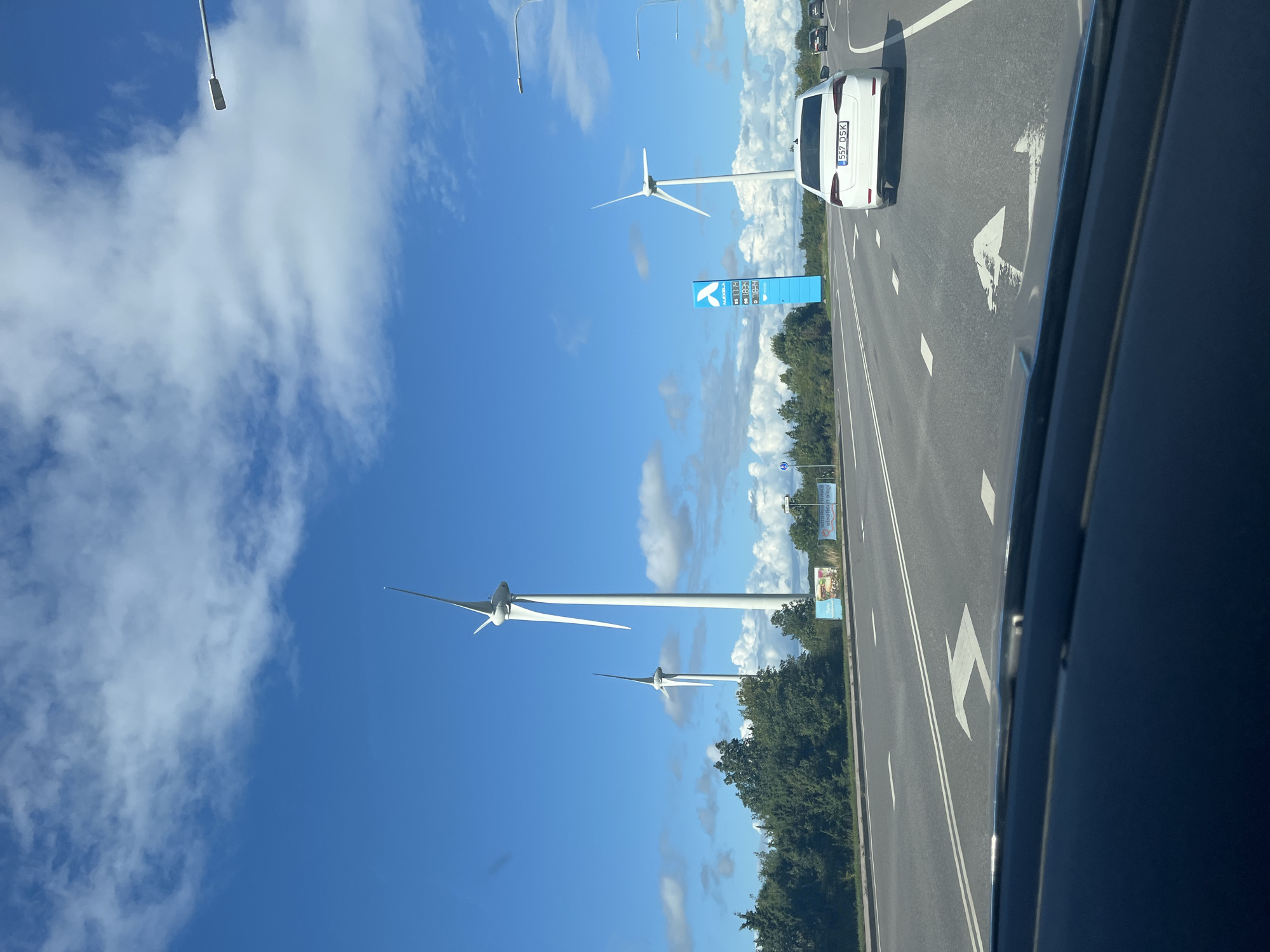 The image shows a bright, sunny day in Virtsu, Estonia. The sky is a vibrant blue with scattered white clouds. In the foreground, a car is driving on a highway. To the right of the highway, a white modern car with the license plate "557 DSK" is visible. Further ahead, a tall, blue sign displays fuel prices and the ALEXELA logo. Behind the sign, a wind turbine stands tall. To the left of the highway, trees line the road, and two more wind turbines are visible in the distance. A few billboards are also present along the roadside. The overall scene depicts a typical roadside view in a coastal town with modern infrastructure.