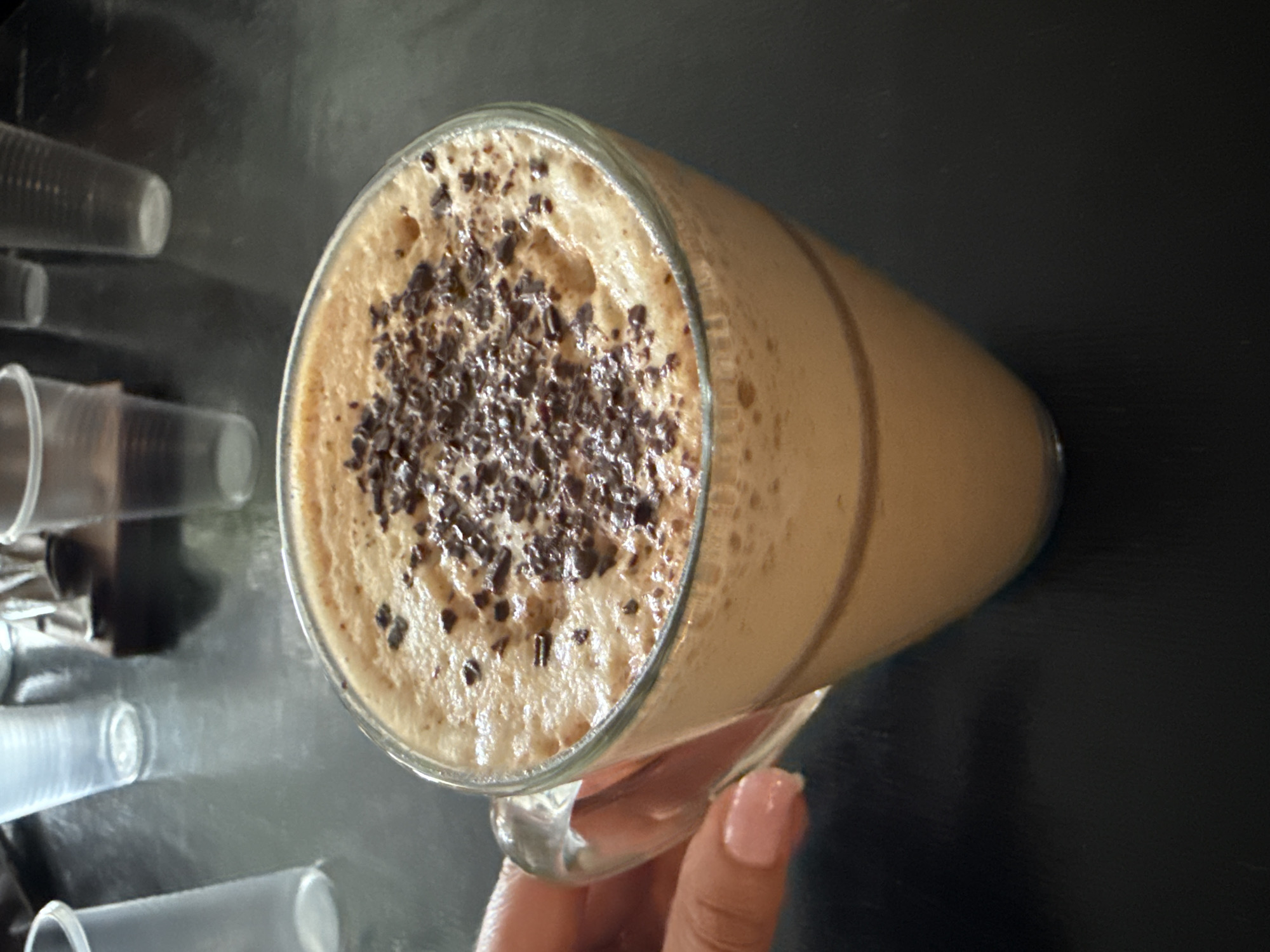 A close-up shot shows a glass of frothy coffee topped with chocolate shavings. The glass is held by a hand with a pink nail, presumably a woman's hand. To the left of the coffee glass, several clear plastic cups are stacked, suggesting a cafe or restaurant setting. The background is a dark, reflective surface. Ust-Koksa, Russia is not evident.