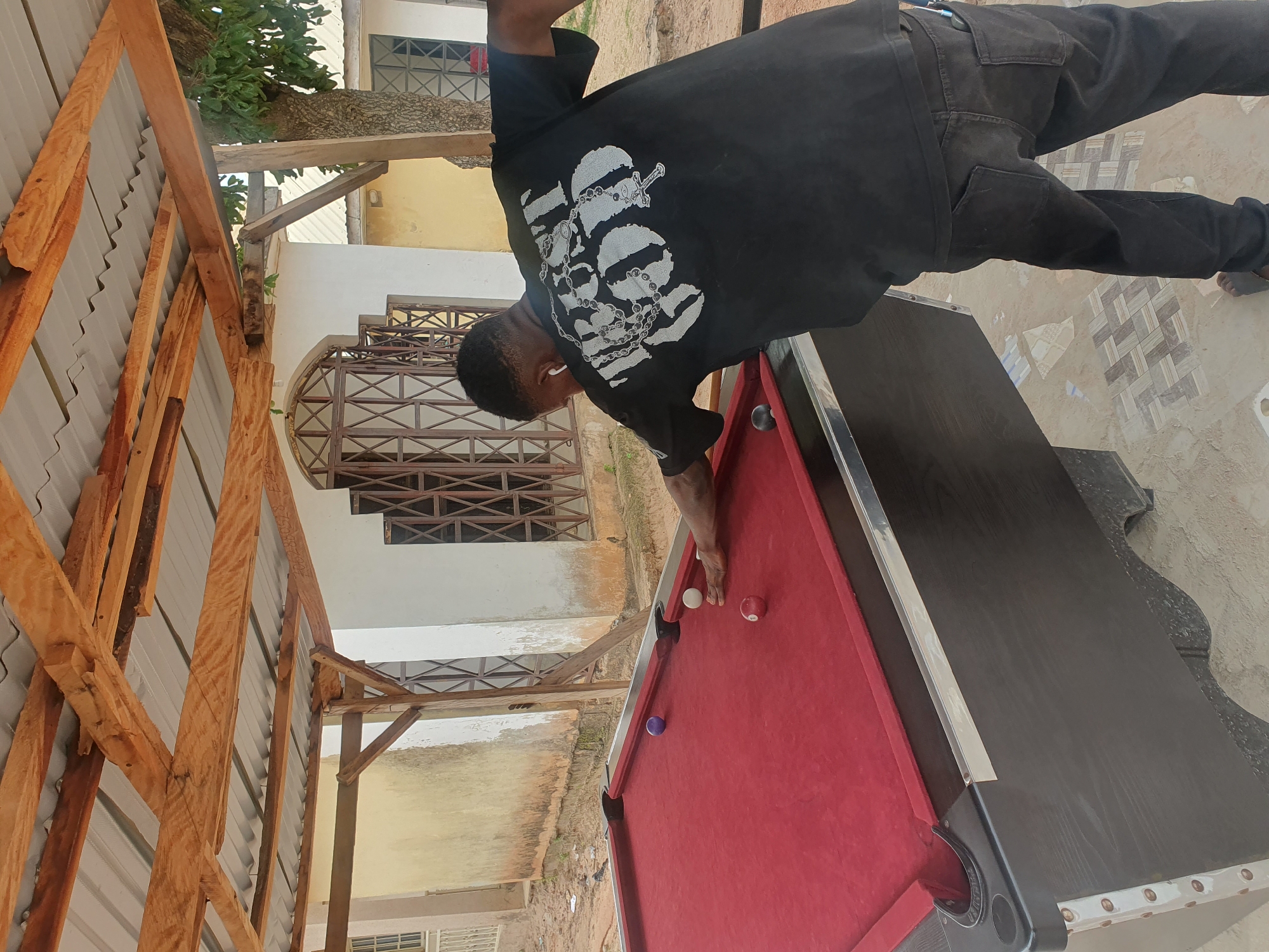 An African man in a black t-shirt and black jeans plays pool at an outdoor table under a wooden, corrugated metal roof. The billiard table is red with black trim. Several billiard balls are on the table, including a purple ball, a white ball, and a red and white striped ball. The man is leaning over the table, his right hand on a cue stick, preparing to strike the white ball. He is wearing white earbuds. The background shows a light-colored building with decorative metal grilles on the windows and a tree. The ground is sandy and tiled in some areas. The scene appears to be in a residential or commercial area in Lokoja, Nigeria.
