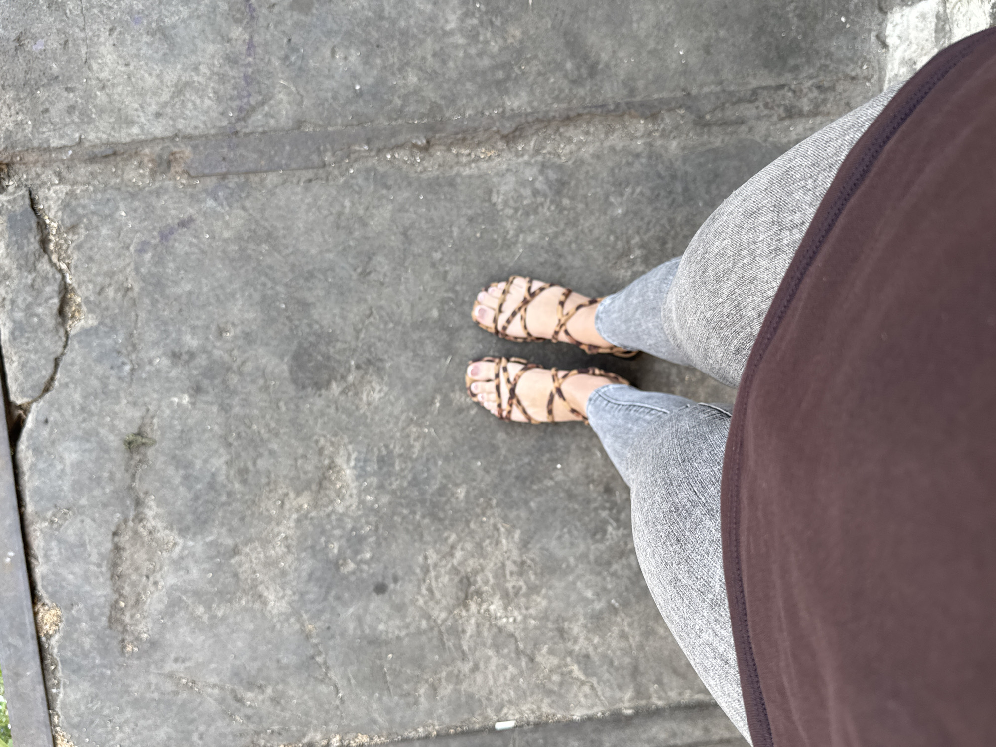 A pair of feet wearing leopard print sandals and grey jeans stand on a cracked concrete surface in the city of Unknown, Unknown. The person is wearing a brown shirt, which is visible at the bottom of the frame. The concrete appears weathered and uneven, with some debris scattered on its surface.