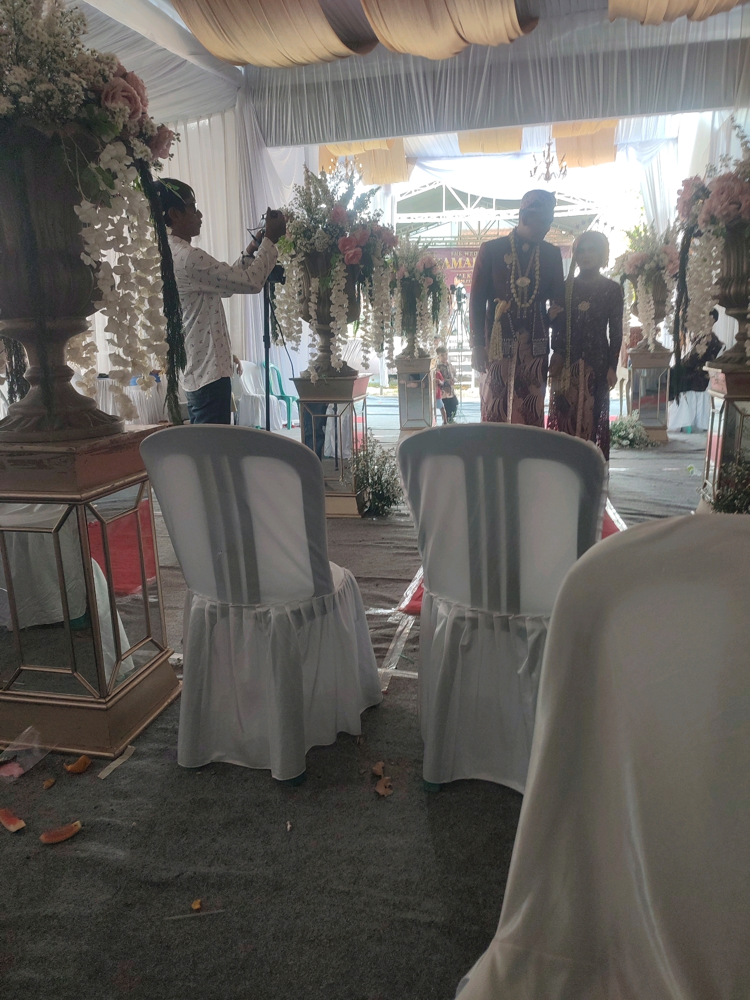 A medium shot captures a wedding ceremony taking place under a draped tent in Kecamatan Haurgeulis, Indonesia. In the foreground, two chairs are draped in white fabric, their backs visible as they face away from the camera. To the left, a person in a white shirt with a patterned design is focused on adjusting a camera on a tripod. Behind them, floral arrangements in large urns adorn the scene, with cascading white flowers and pink roses. In the middle ground, a couple, presumably the bride and groom, stand dressed in traditional attire. The groom wears a maroon jacket and a patterned sarong, adorned with necklaces, while the bride is in a matching patterned dress. Behind them, the tent's draped fabric ceiling creates a warm, diffused lighting. Scattered on the carpeted ground are fallen flower petals and debris. The overall impression is one of a formal celebration, with attention to traditional customs and decorations.