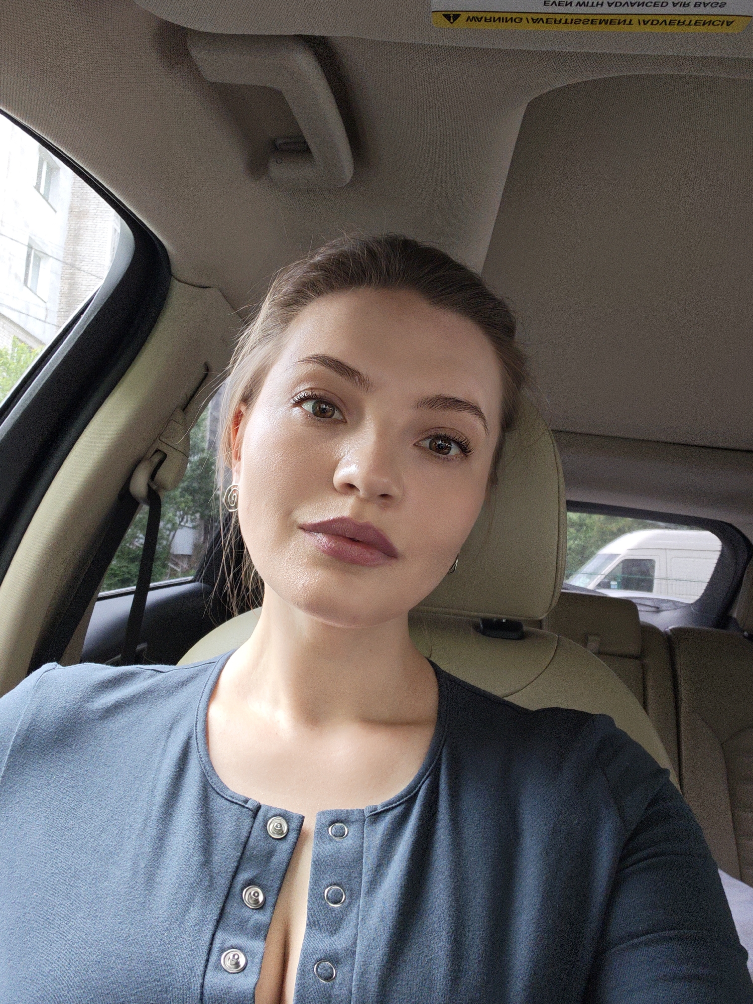 A woman is seated in the driver's seat of a car. She has light skin, brown hair, and is wearing a blue button-down shirt. The car's interior is light beige. Outside the car window, a building and trees are visible. The text on the car's sun visor is reflected and unreadable. The location cannot be determined.