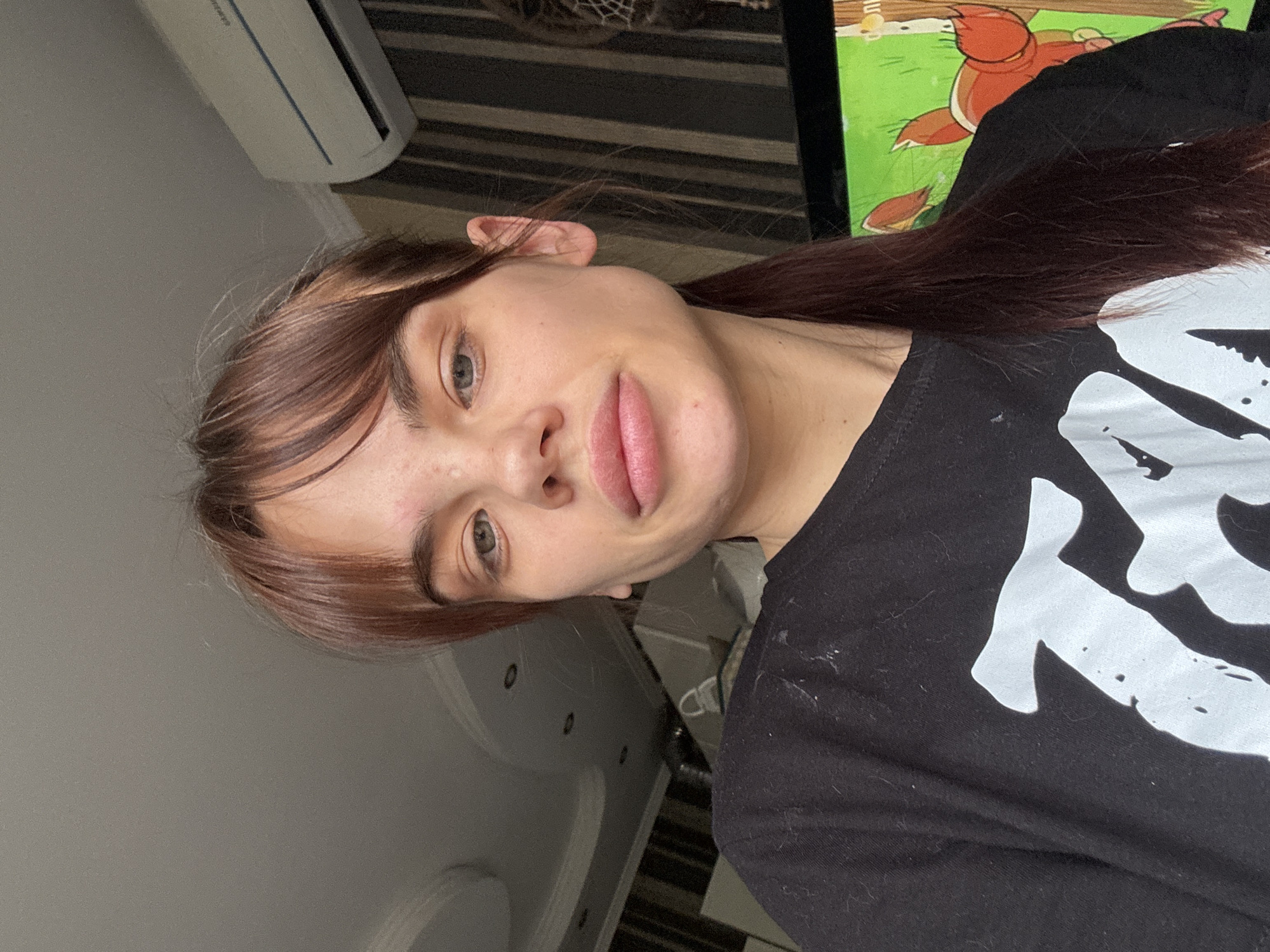 A young woman with light skin, light brown hair, and gray eyes is looking directly at the camera. She has full lips and some freckles on her face. She's wearing a black t-shirt with a white graphic design on it. The background includes a striped wall and what appears to be a television displaying a cartoon image. Part of an air conditioning unit is visible in the upper left. The picture appears to be taken indoors. There is no information in the image to confirm this is Rostov-on-Don, Russia.