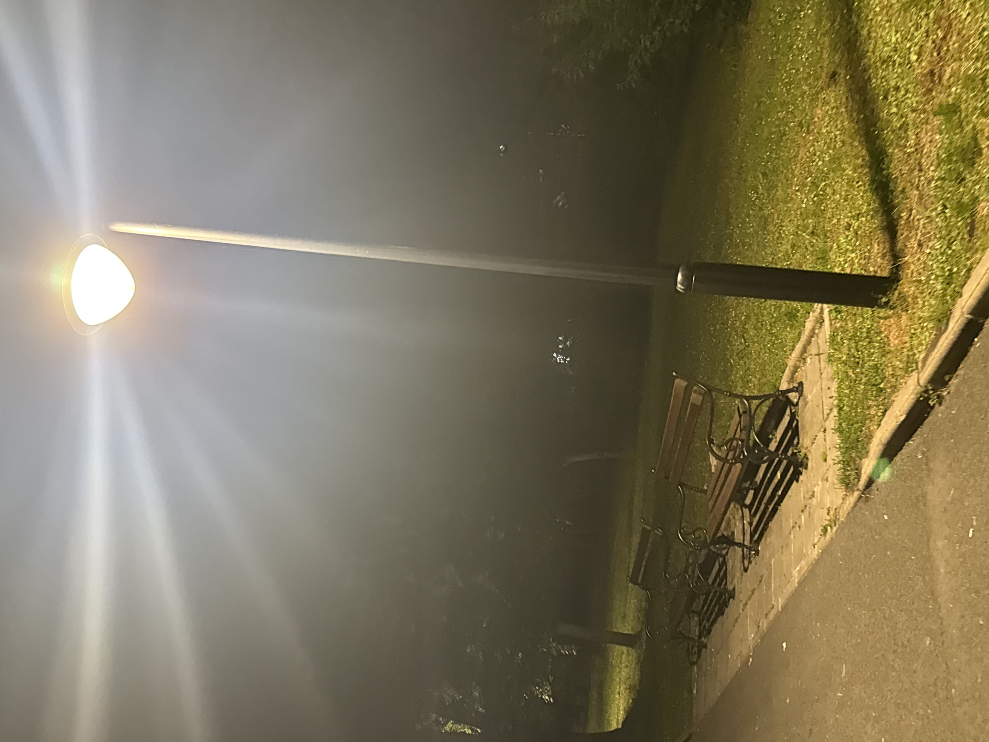 The image depicts an outdoor scene at night, illuminated by a street lamp. The lamp, positioned to the right of the frame, has a bright, glowing bulb. Its light casts a strong beam across the upper portion of the image, creating visible rays. Below the lamp, a dark, slender pole extends downwards.
In the foreground, a paved pathway runs diagonally from the bottom left corner towards the middle of the image. Adjacent to the pathway, on the right, is a strip of green grass. Several park benches with dark metal frames and wooden slats are situated on a paved area next to the grass, further to the right. Trees are visible in the background, their forms obscured by the darkness and the lighting conditions. The overall atmosphere is somewhat hazy, suggesting either fog or mist.
The location is identified as Burgas, Bulgaria.