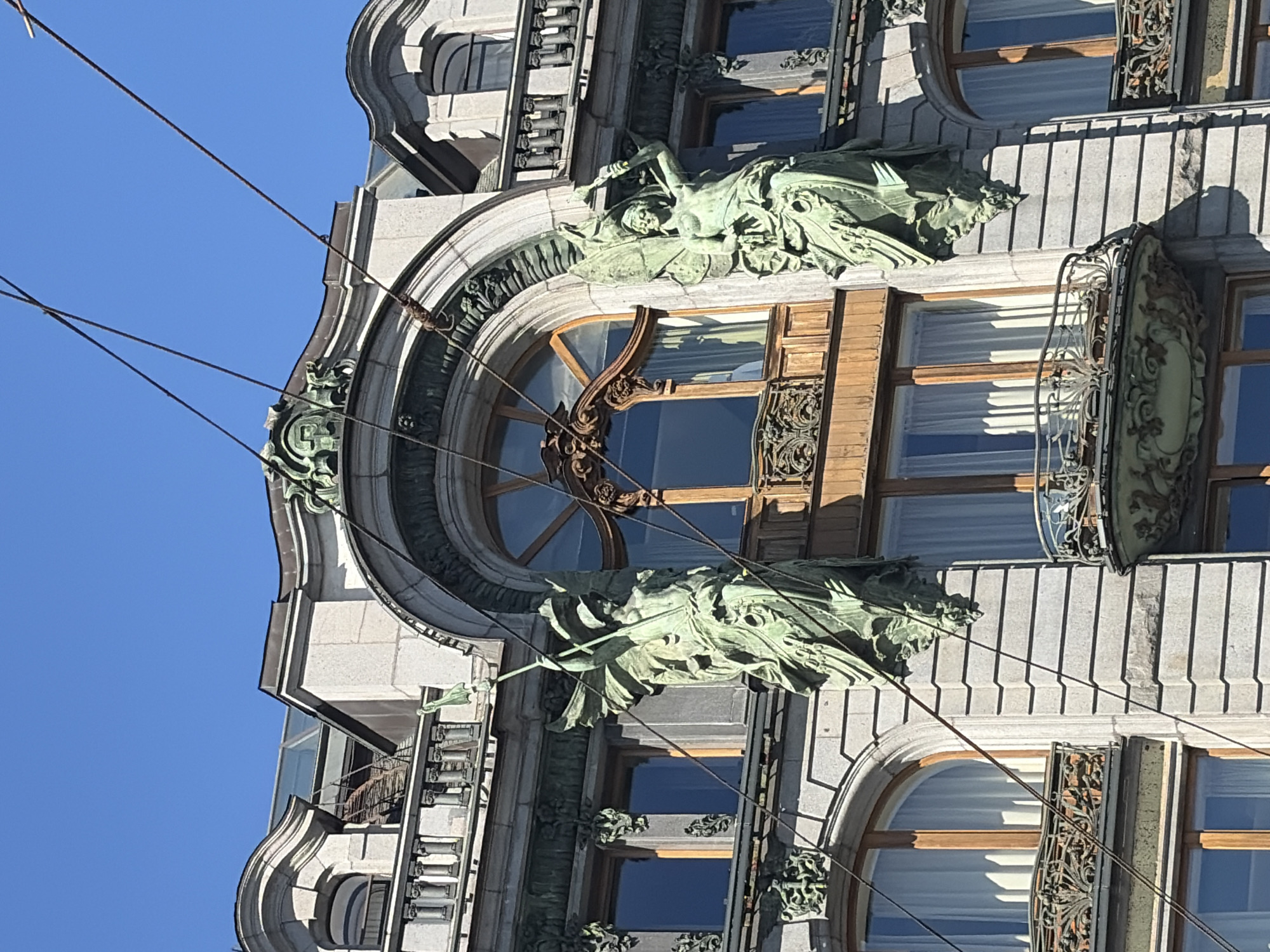 This image presents an upward-looking view of a highly ornate architectural facade, characteristic of historical European cities like Saint Petersburg, Russia, under a clear, bright blue sky. The building appears to be constructed from light-colored stone or stucco, featuring intricate carvings and decorative elements that suggest a blend of classical and possibly Art Nouveau styles.
The central focus is a grand arched window, framed by a wooden structure and adorned with elegant metal grille work, which reflects the clear sky. Flanking this prominent window are two large, verdigris-patinated bronze or copper statues or high reliefs. These figures, possibly allegorical, are depicted in flowing drapery and classical poses, adding a dramatic and artistic dimension to the facade. The figure on the right (as seen in the original, rotated image) appears to hold a staff or spear.
Below the main arched window, other rectangular windows are visible, some featuring decorative, scrolled metal balconies and railings. The strong sunlight casts sharp, well-defined shadows across the building's textured surfaces, accentuating the depth and detail of the sculptures and architectural features.
Several dark, thin wires, likely overhead power lines for urban transport like trams or trolleybuses, traverse the foreground, partially intersecting the building's upper sections. The overall impression is one of grandeur and historical artistry. The bright, clear sky and distinct shadows indicate a sunny day, likely mid-morning or afternoon. No specific human activity is depicted, nor is any clear, legible text visible on the facade.