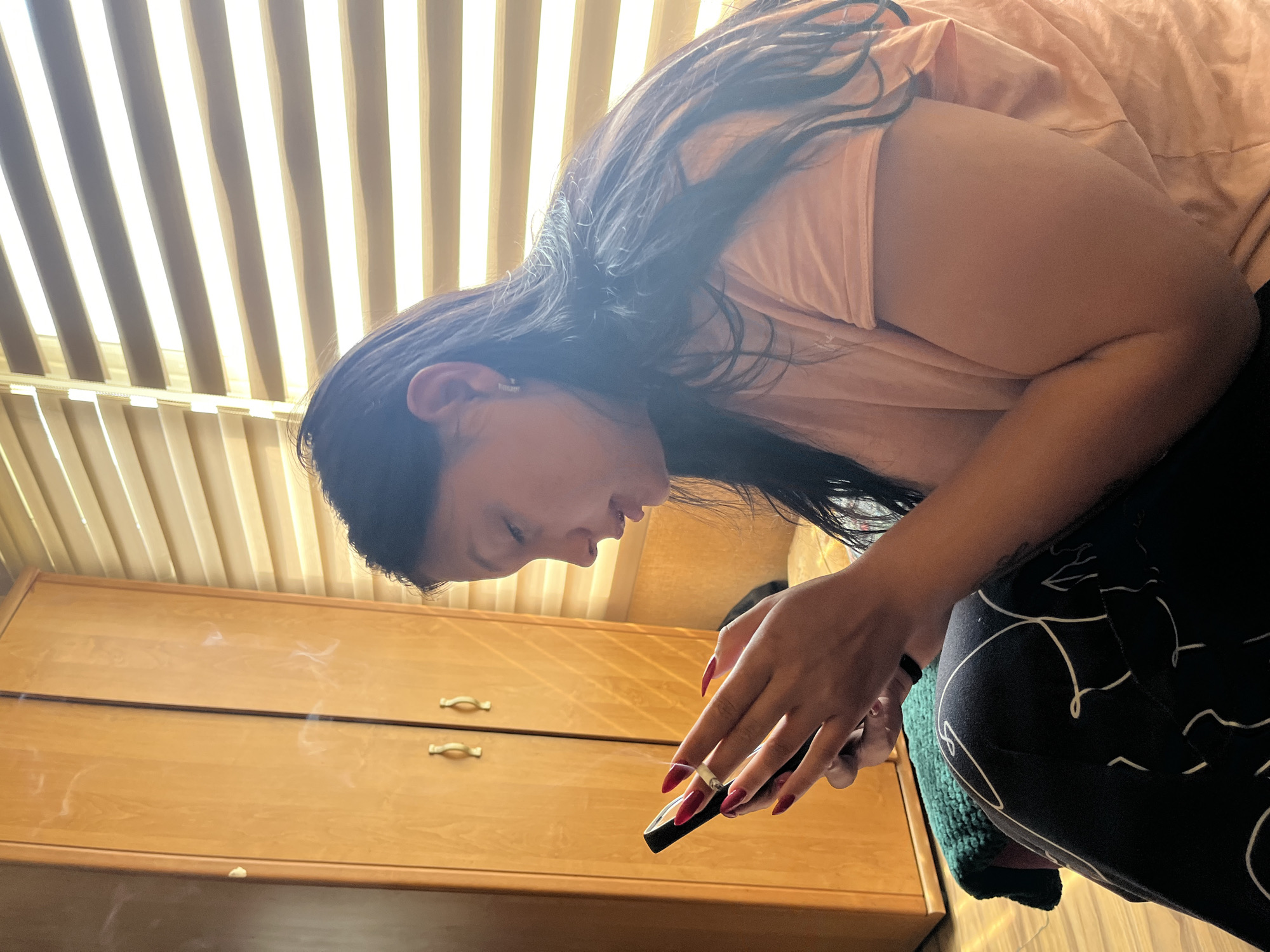 A woman with long, dark hair is bending forward and looking at a black mobile phone she is holding. She has red nail polish and a ring on her left hand. She is wearing a light pink shirt and black and white patterned pants. The background includes a light brown wooden dresser with two drawers and metal pulls, and vertical blinds. It is impossible to determine the location in the city of Sofia, Bulgaria, based on the image.