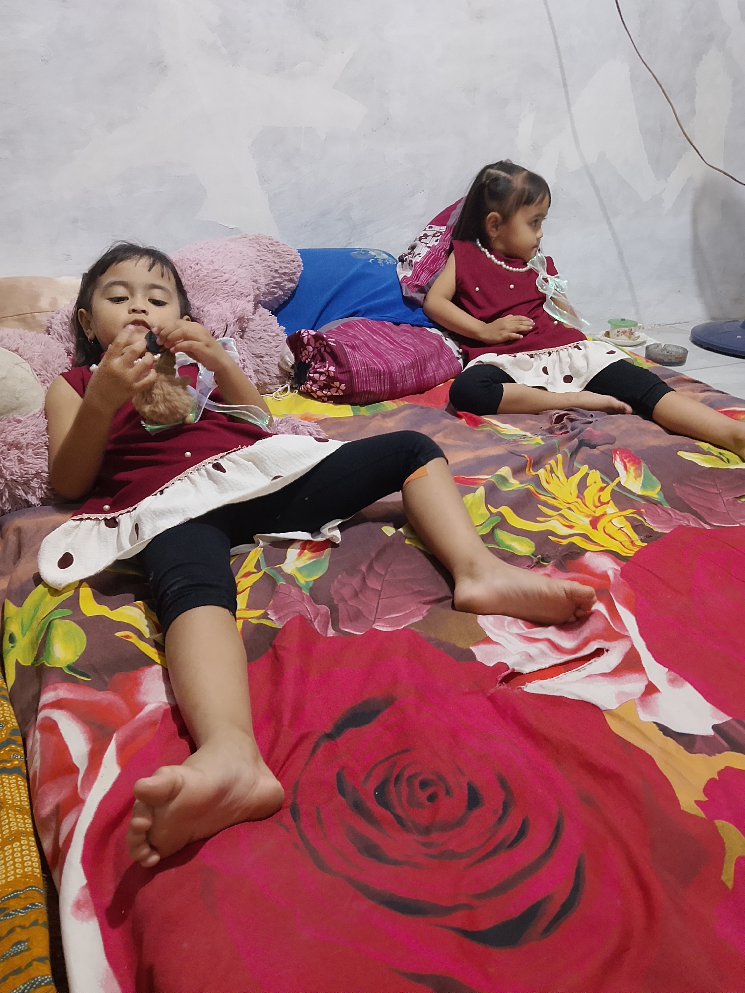 Two young girls are lying on a bed with a floral comforter in a room. The girl in the foreground, on the left, is wearing a dark red sleeveless dress with a white ruffled hem adorned with brown polka dots and a white ribbon tied to her neck. She has black leggings on and is holding a small brown stuffed toy. Her legs are extended, and her left foot is visible. The girl in the background, on the right, is wearing a similar dark red sleeveless dress with a white skirt decorated with brown polka dots. She has a pearl necklace around her neck. She is looking to her right. In the background, a blue pillow and a pink furry pillow are visible. The wall behind the bed is light gray with some textured paint. A brown electrical cord runs from the top right of the frame towards the right. A small green cup on a saucer and an ashtray are on the bed near the girl in the background. The room appears to be dimly lit. The location of this scene is the city of Tanggungan, Indonesia.