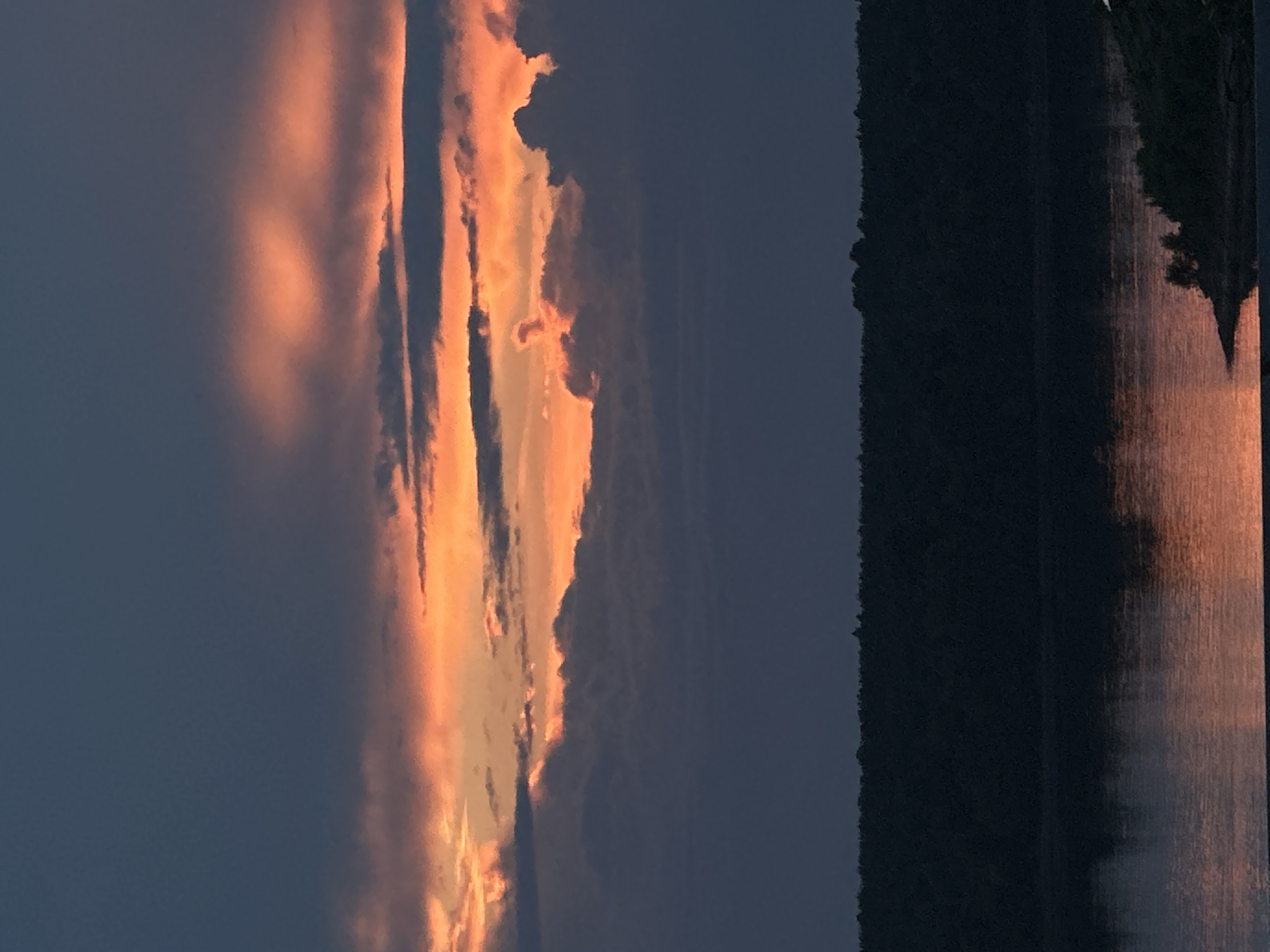 The image depicts a sunset over a body of water, likely the Kama River, near the city of Chaikovsky, Russia. The sky is filled with dramatic clouds, illuminated by the setting sun with vibrant orange and pink hues. These colors are reflected on the calm surface of the water, creating a mirror image of the fiery sky. In the foreground, a dark, tree-covered shoreline stretches across the horizon, silhouetted against the illuminated sky. The overall scene is serene and visually striking, characteristic of a natural landscape at dusk. There are no people or man-made objects visible in the image.