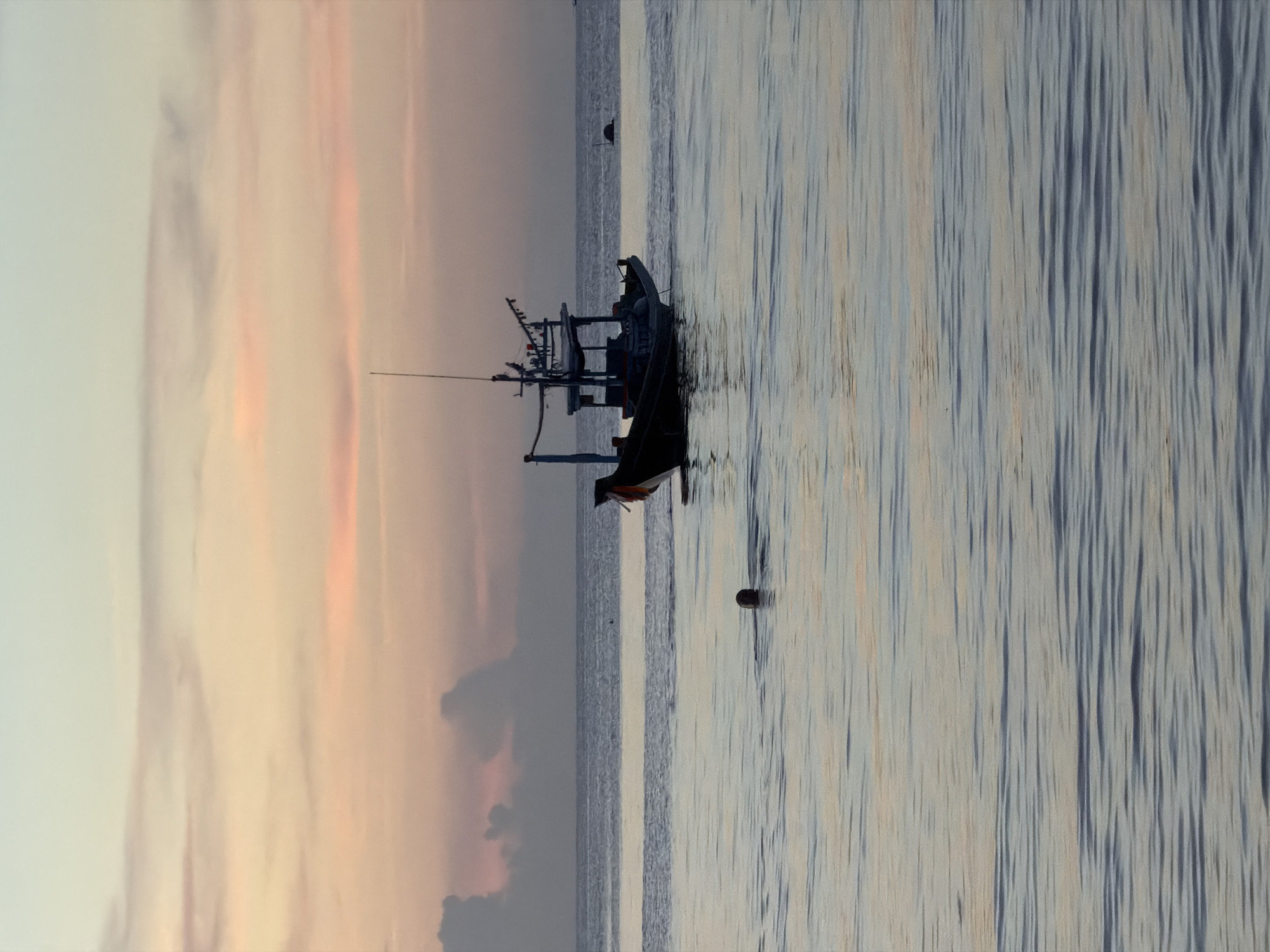 The image captures a serene seascape at dusk, likely on Koh Phangan, Thailand. The sky displays a gradient of soft pastel hues, with streaks of pink and peach blending into a pale blue, characteristic of a tropical sunset or sunrise. A solitary person is visible swimming in the calm ocean, their head just above the water. Further out, a dark fishing boat with a tall mast is anchored or slowly moving on the water. The ocean itself is tranquil, with gentle ripples reflecting the colors of the sky. The overall atmosphere is one of peaceful solitude and natural beauty.