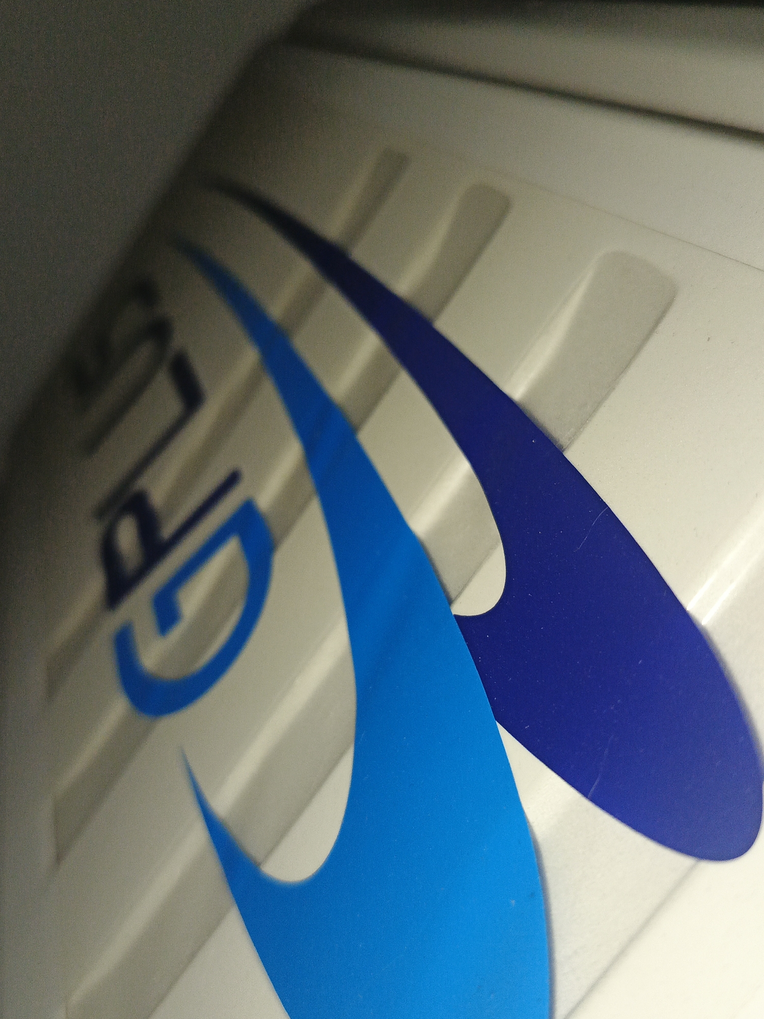 The image shows a close-up of a white object with the partially visible words "GP PLUS" in blue on the left. Two curved shapes, one in light blue and another in dark blue, are prominently featured. The object has a grooved surface with parallel indentations. The lighting is uneven, with the top left area appearing darker. The location and presence of people, specific subjects, actions, and interactions cannot be determined from the image alone. Therefore, I cannot confirm it is in Los Robles, Venezuela.